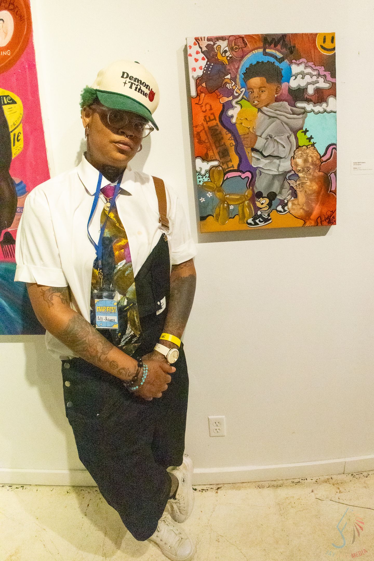 Young artist poses in white shirt, tie, and cap beside colorful pop art paintings at an art gallery exhibition.