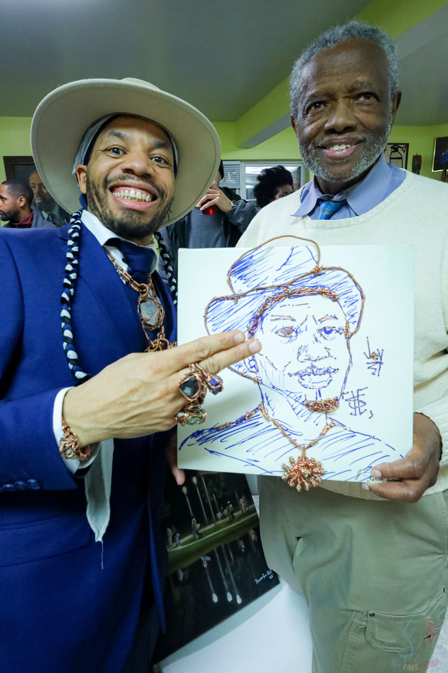 Two men smiling and holding a hand-drawn portrait sketch at an indoor event, one wearing a blue suit and hat.
