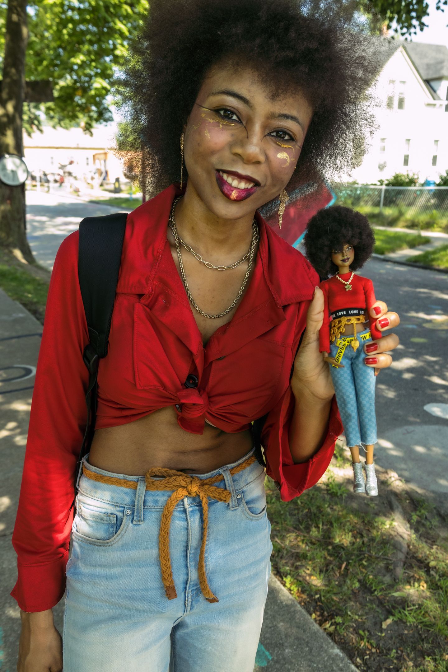 A smiling woman with an afro holds a matching doll dressed in red at an outdoor community festival.