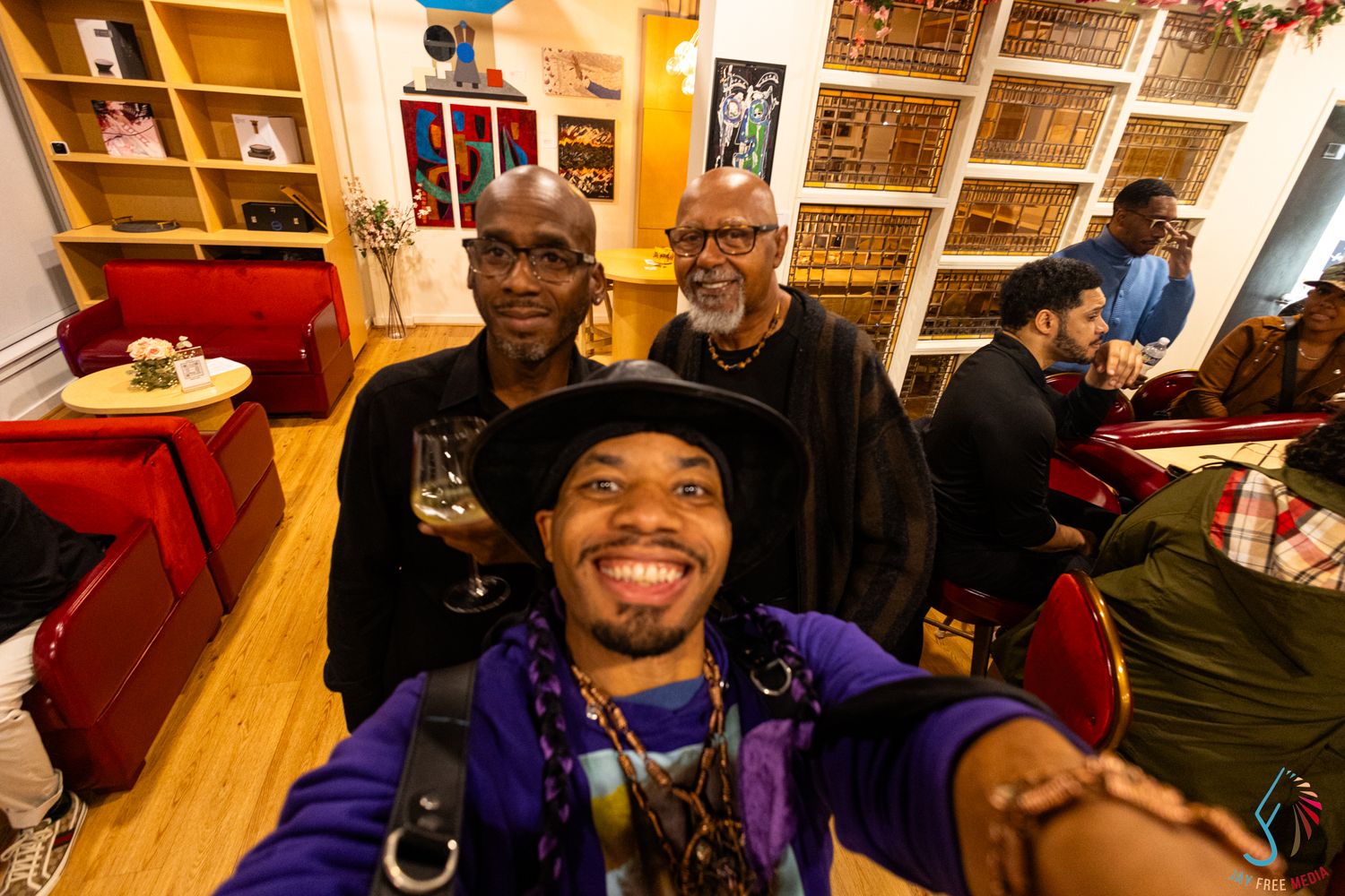 Three men smiling for a selfie inside a cozy room with shelves and warm lighting.