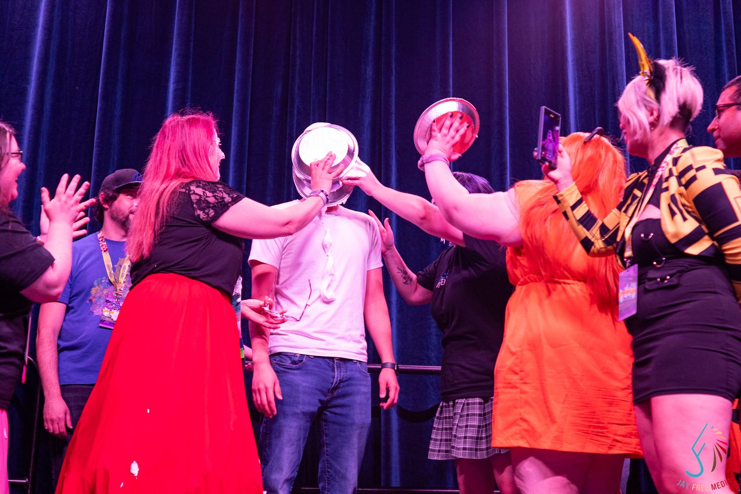 Group of cosplayers on stage placing a shiny helmet on a mans head at a convention event.