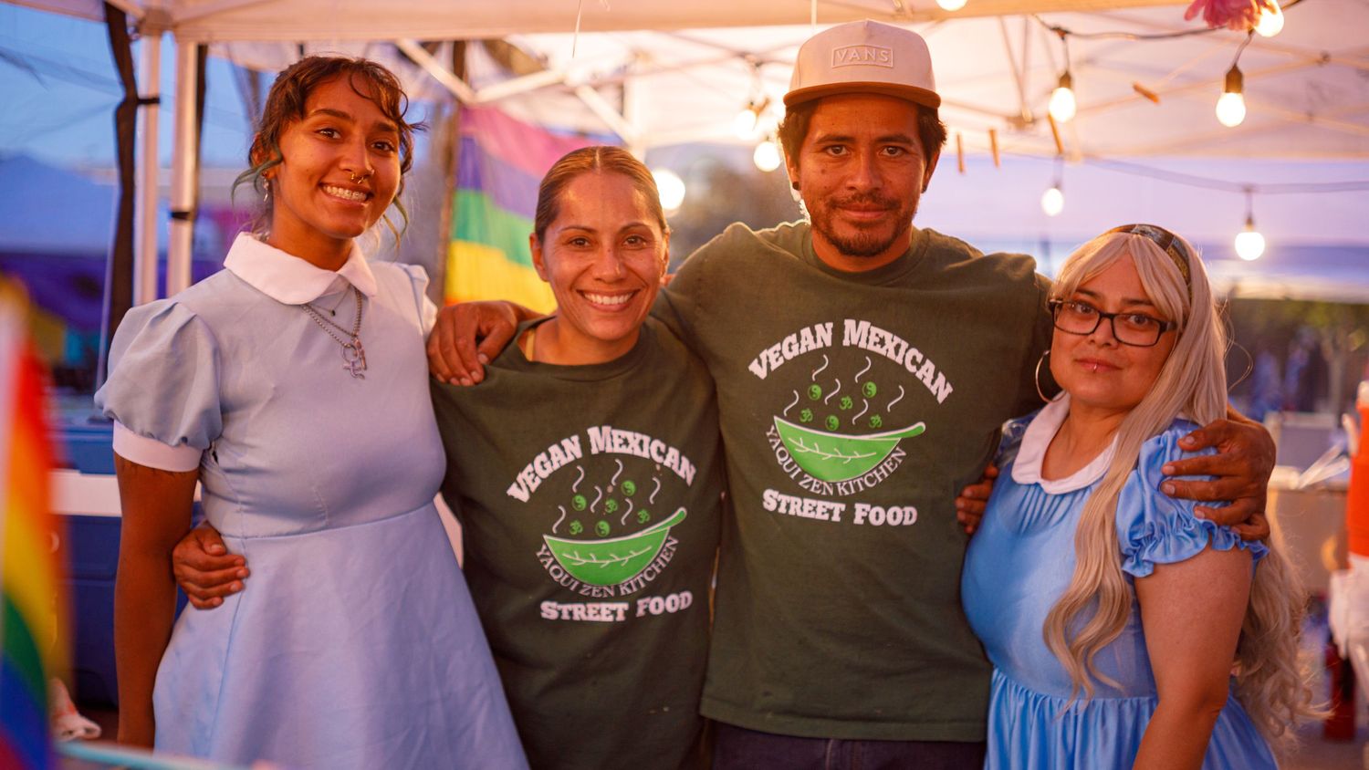 Four people in vegan Mexican street food shirts smile together at outdoor festival booth.