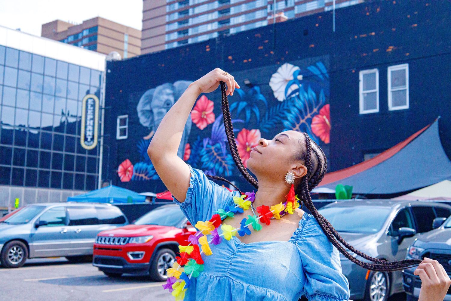 Young Black woman in blue dress playfully holds colorful braided hair up near a tropical hibiscus street mural.