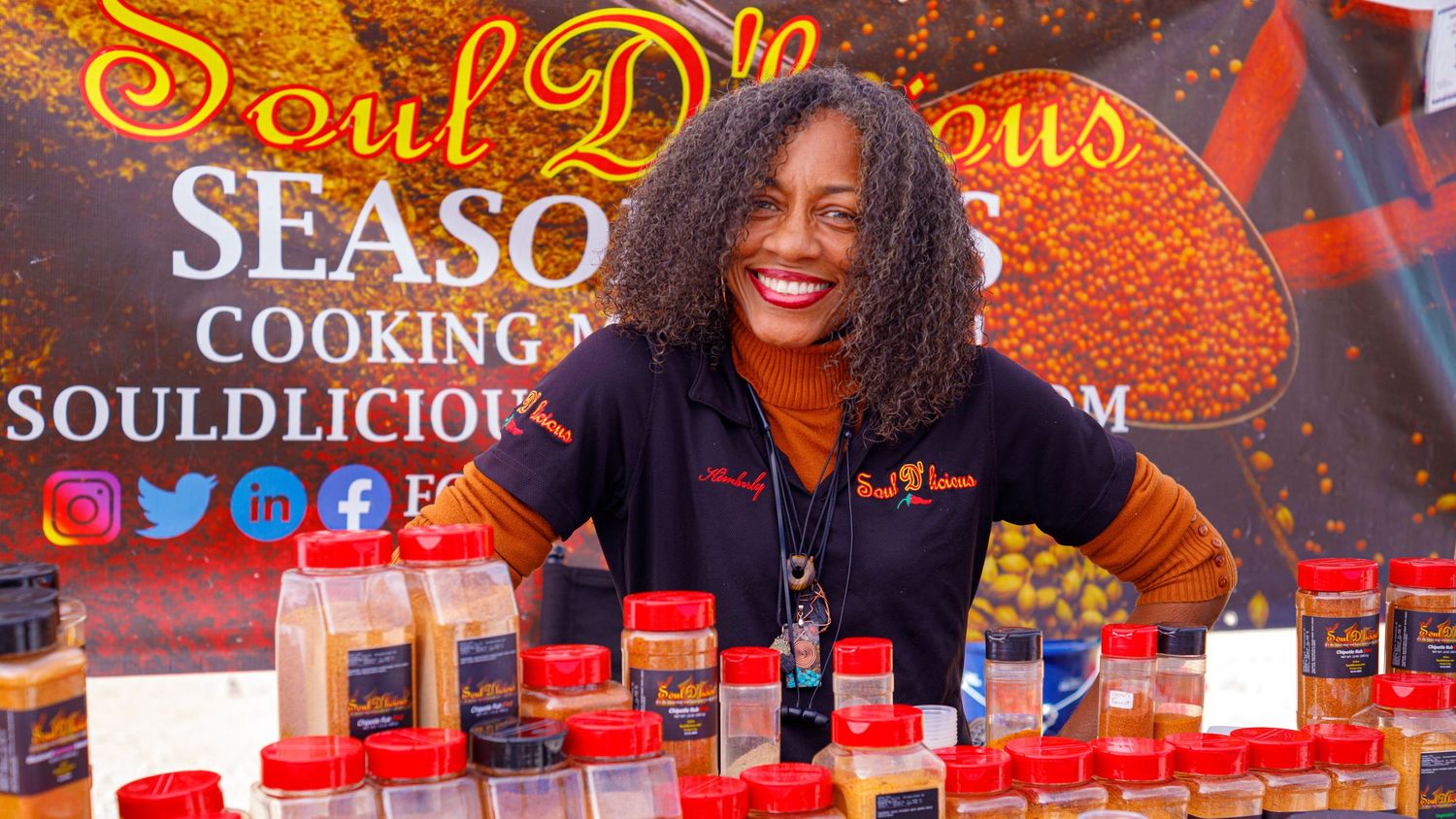 Woman smiling behind a table of Soul Dlicious Seasonings products at an outdoor market.