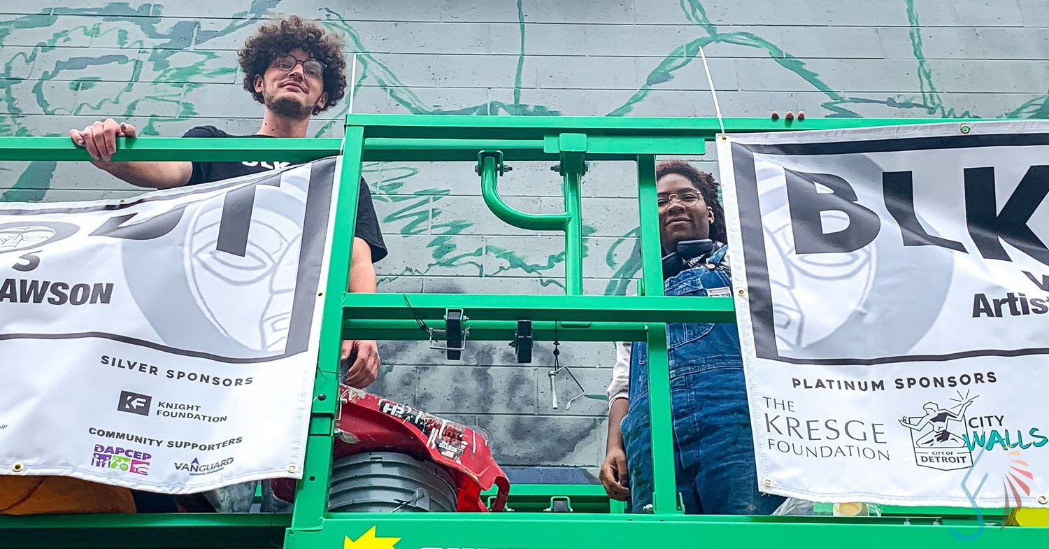 Two artists stand on a green lift platform with BLK and Kresge Foundation sponsor banners.