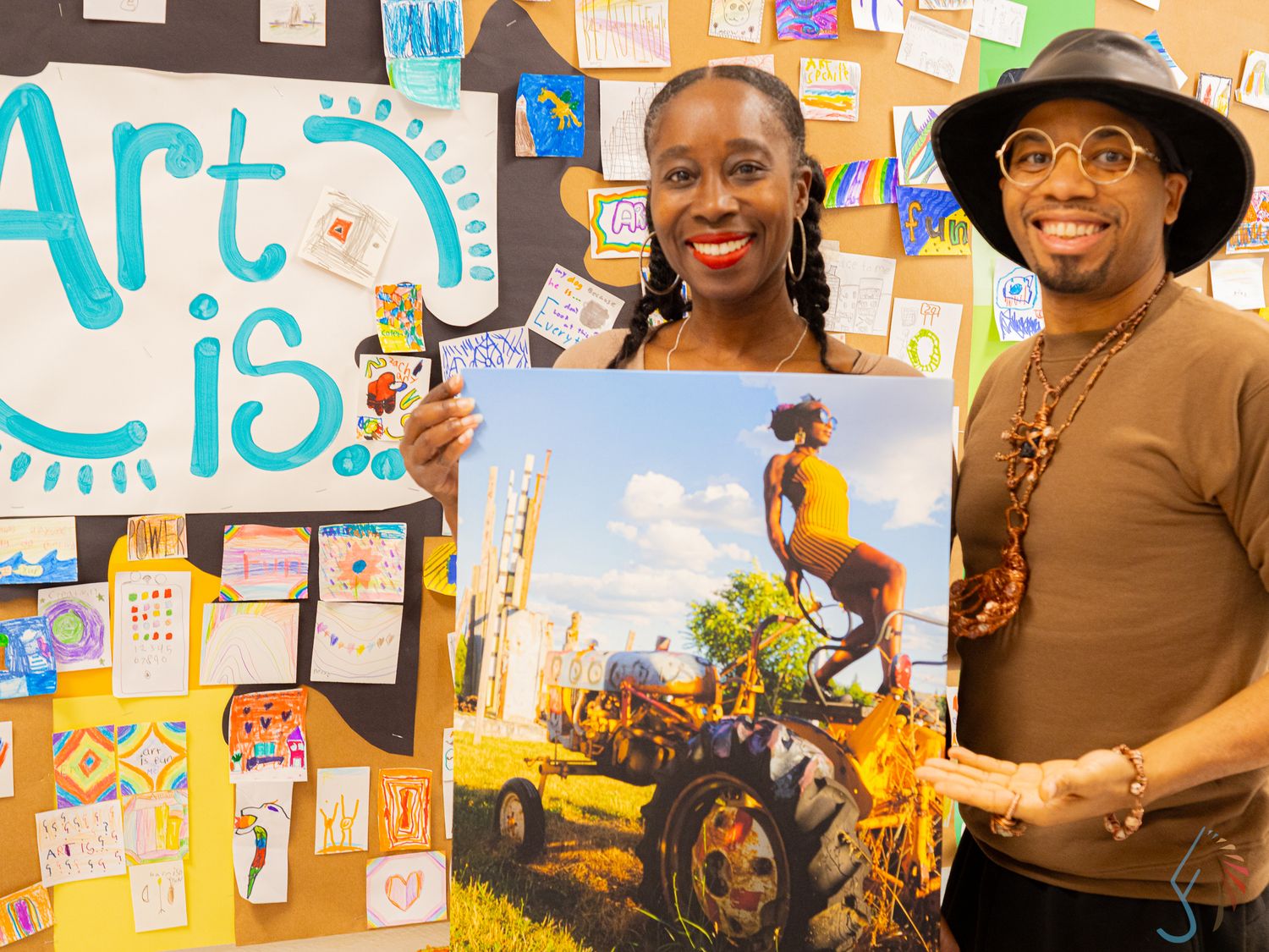 Two people proudly display colorful artwork in front of a bulletin board with an Art Is sign.