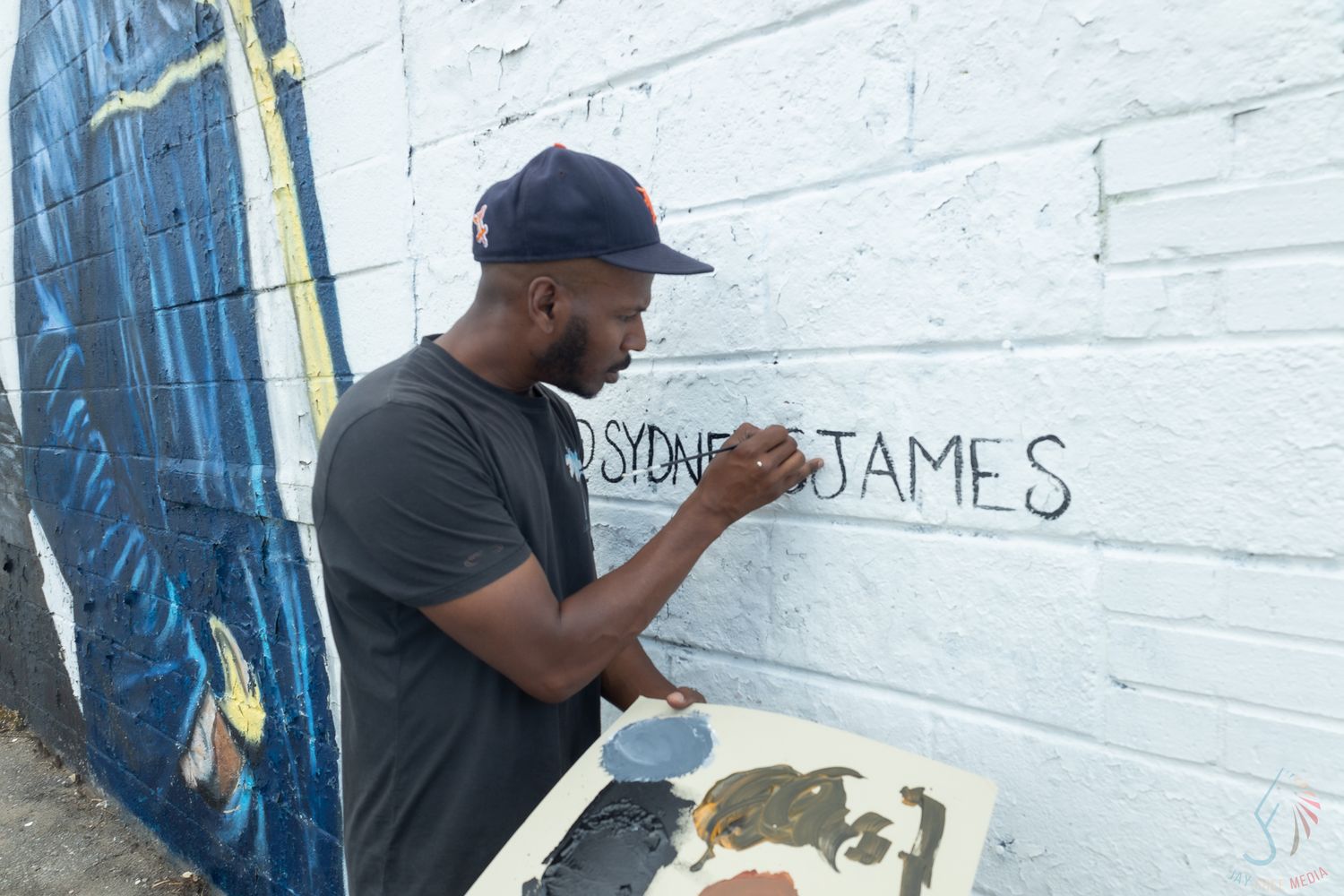 Artist Sydney James painting their name on a white brick wall mural outdoors.