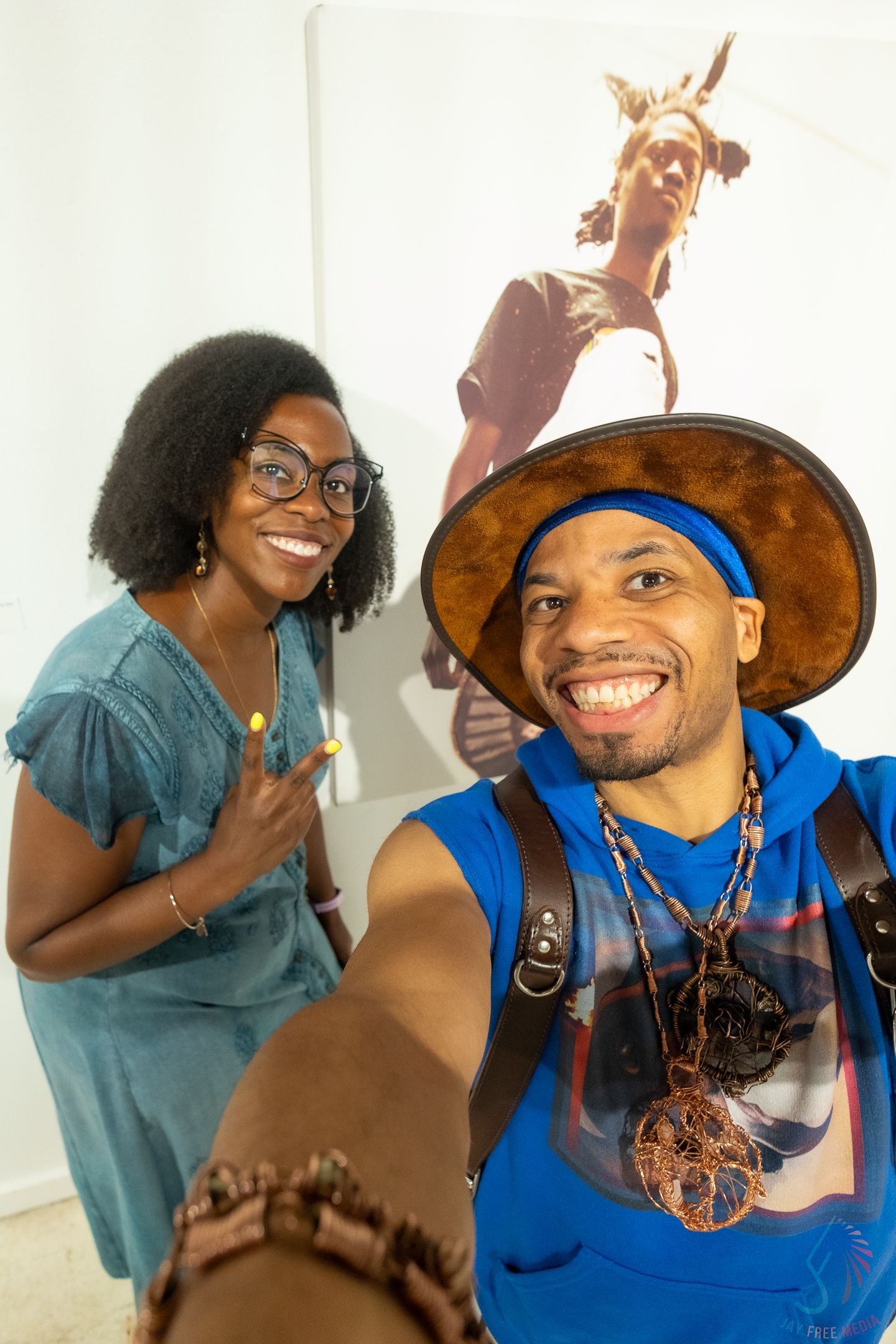 Two smiling Black friends take a selfie at an art gallery, man wearing blue hoodie and brown hat with necklaces.