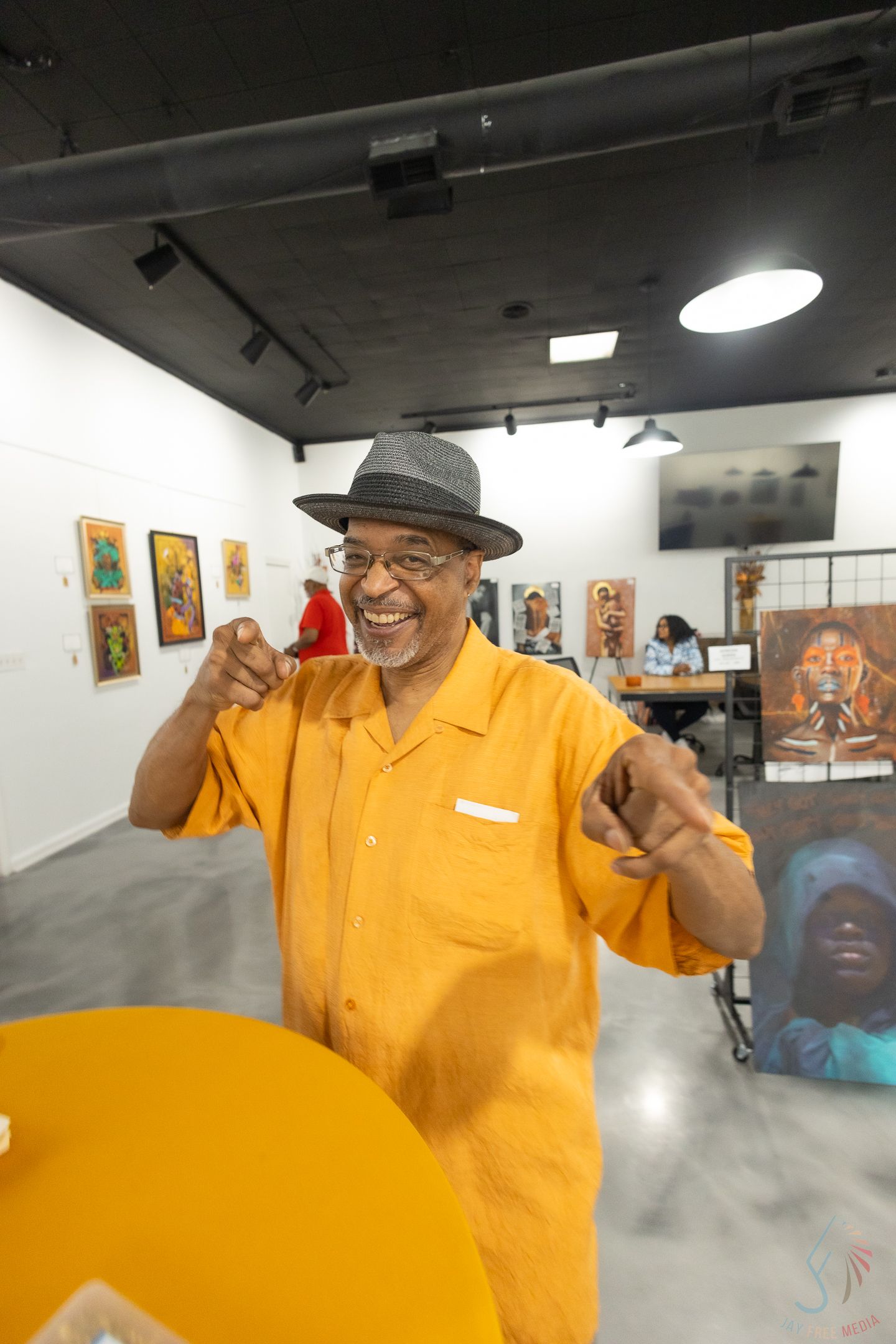 Smiling man in orange shirt and gray fedora hat gestures enthusiastically inside an art gallery.