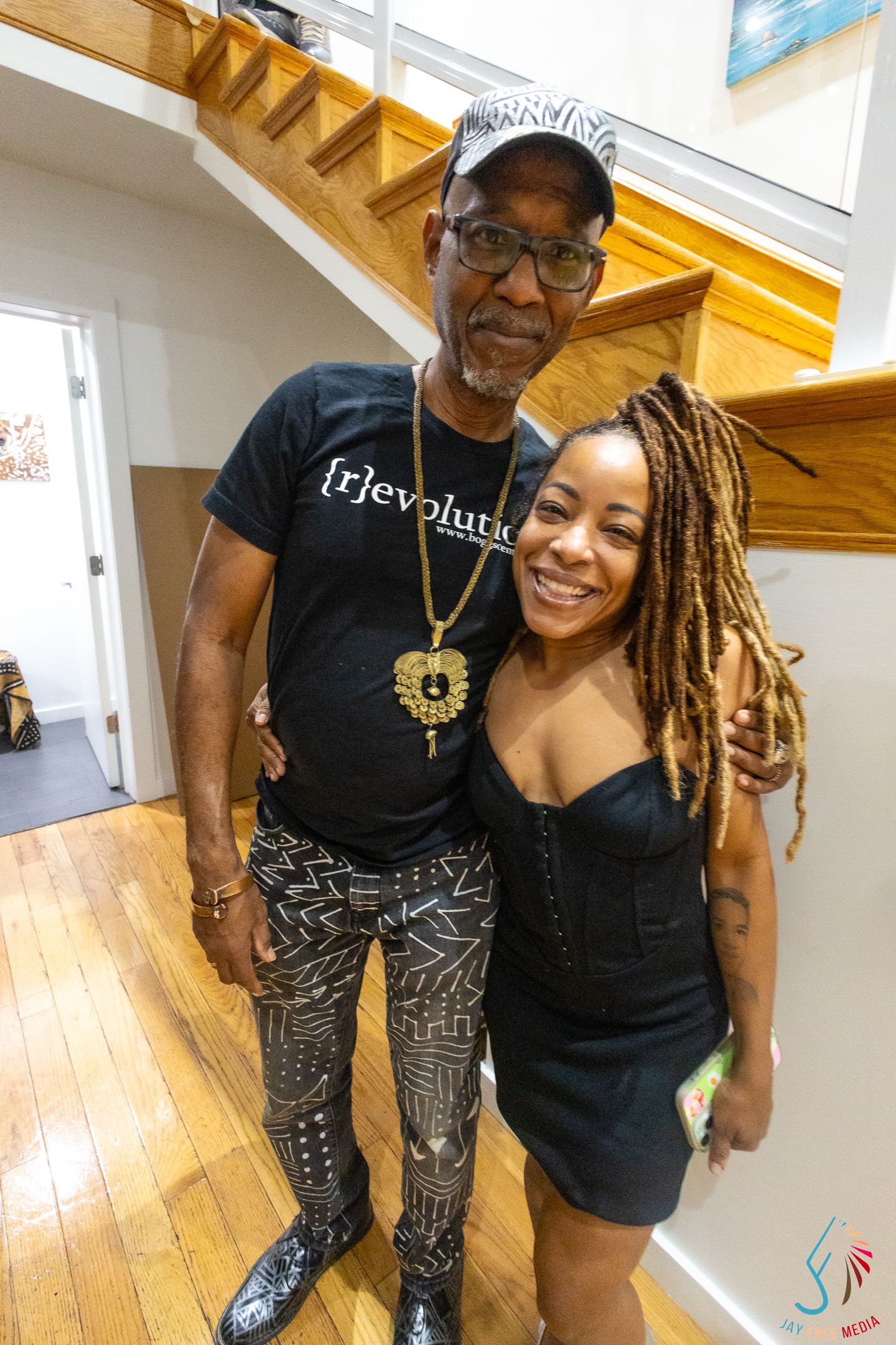 Two people smiling and posing together indoors near a staircase, man wearing a black frjevolution t-shirt and patterned pants.