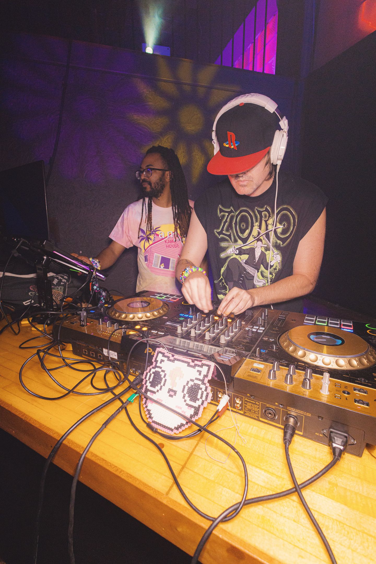 Two DJs performing at a nightclub, one wearing headphones and a PlayStation cap, mixing tracks on Pioneer DJ equipment.