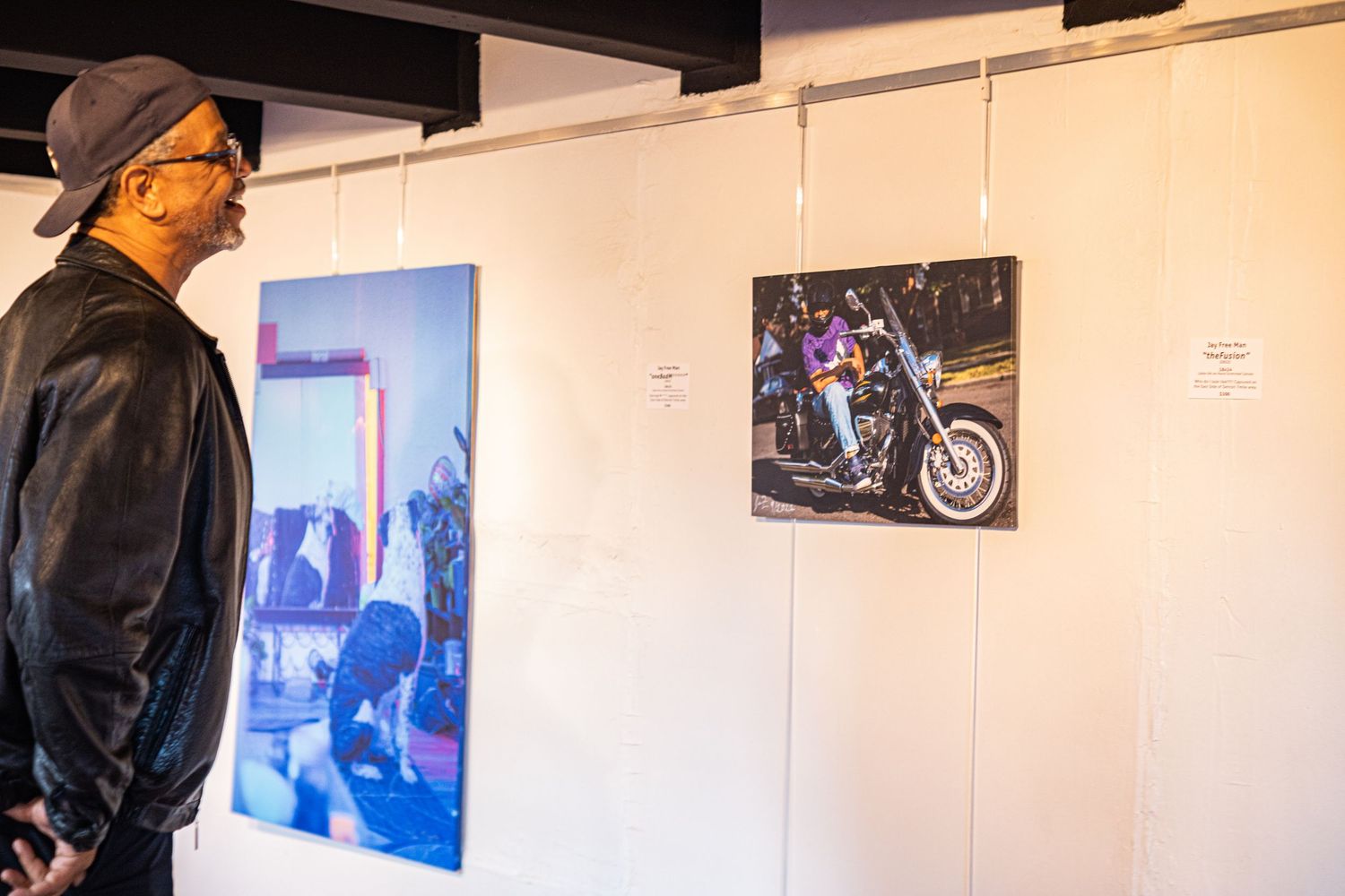 Man in black jacket views motorcycle and street photography prints displayed on white gallery walls.