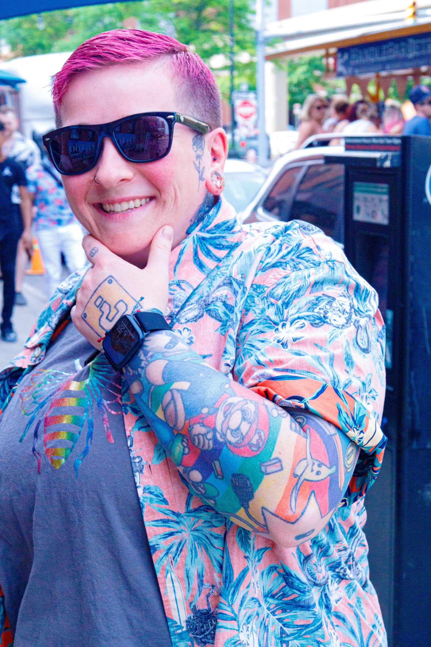 Person with pink hair, sunglasses, floral shirt, and tattoos smiling outdoors on a busy street.