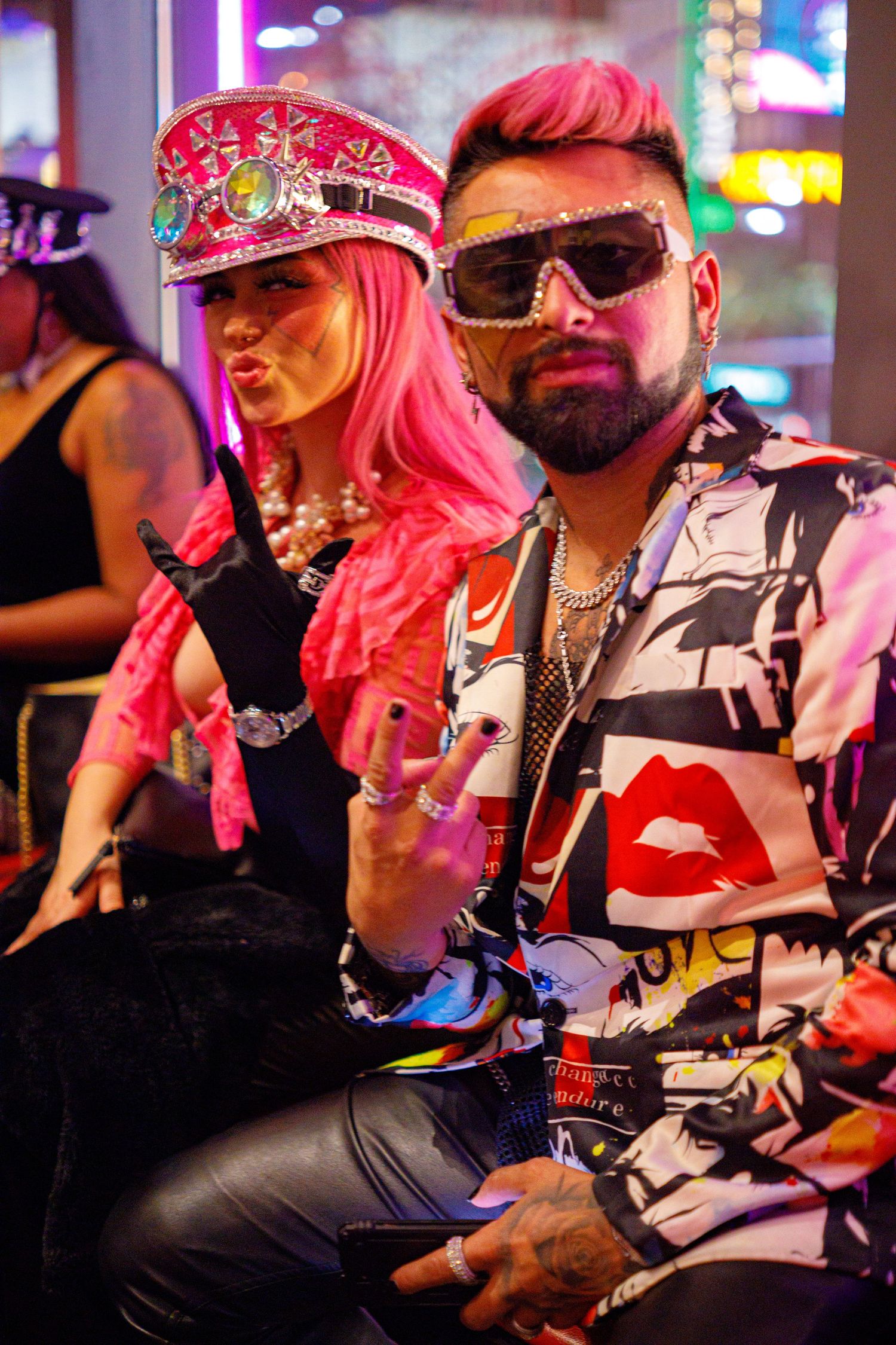 Man in bold printed shirt and futuristic sunglasses poses with peace sign beside woman in pink hair and sequined hat.
