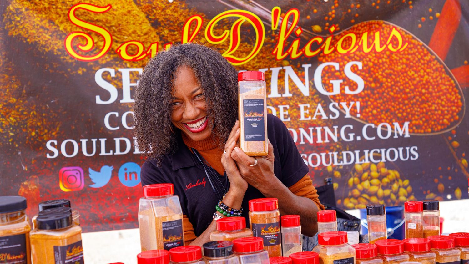 Woman smiling and holding seasoning bottle at Soul D'licious Seasonings vendor booth with spice products displayed.