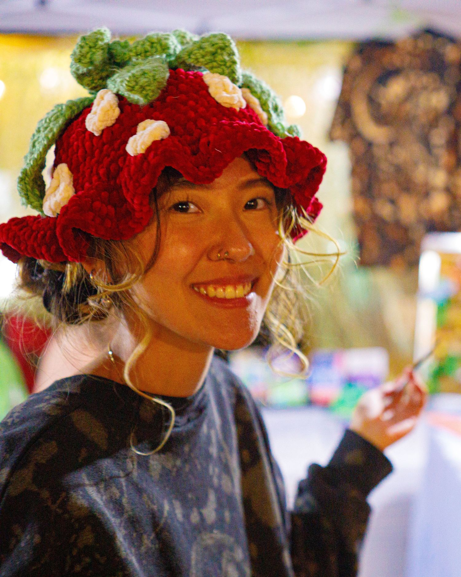 Smiling woman wearing a handmade red strawberry crochet bucket hat at a craft market.