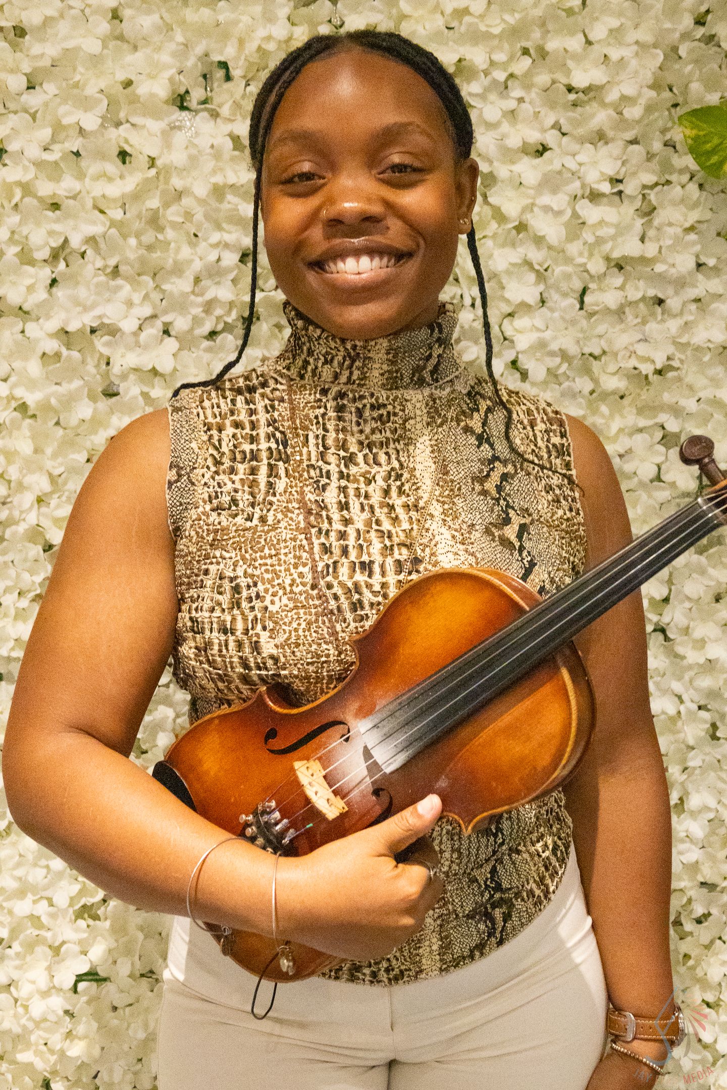 Smiling young woman holding a violin in front of a white floral wall backdrop.