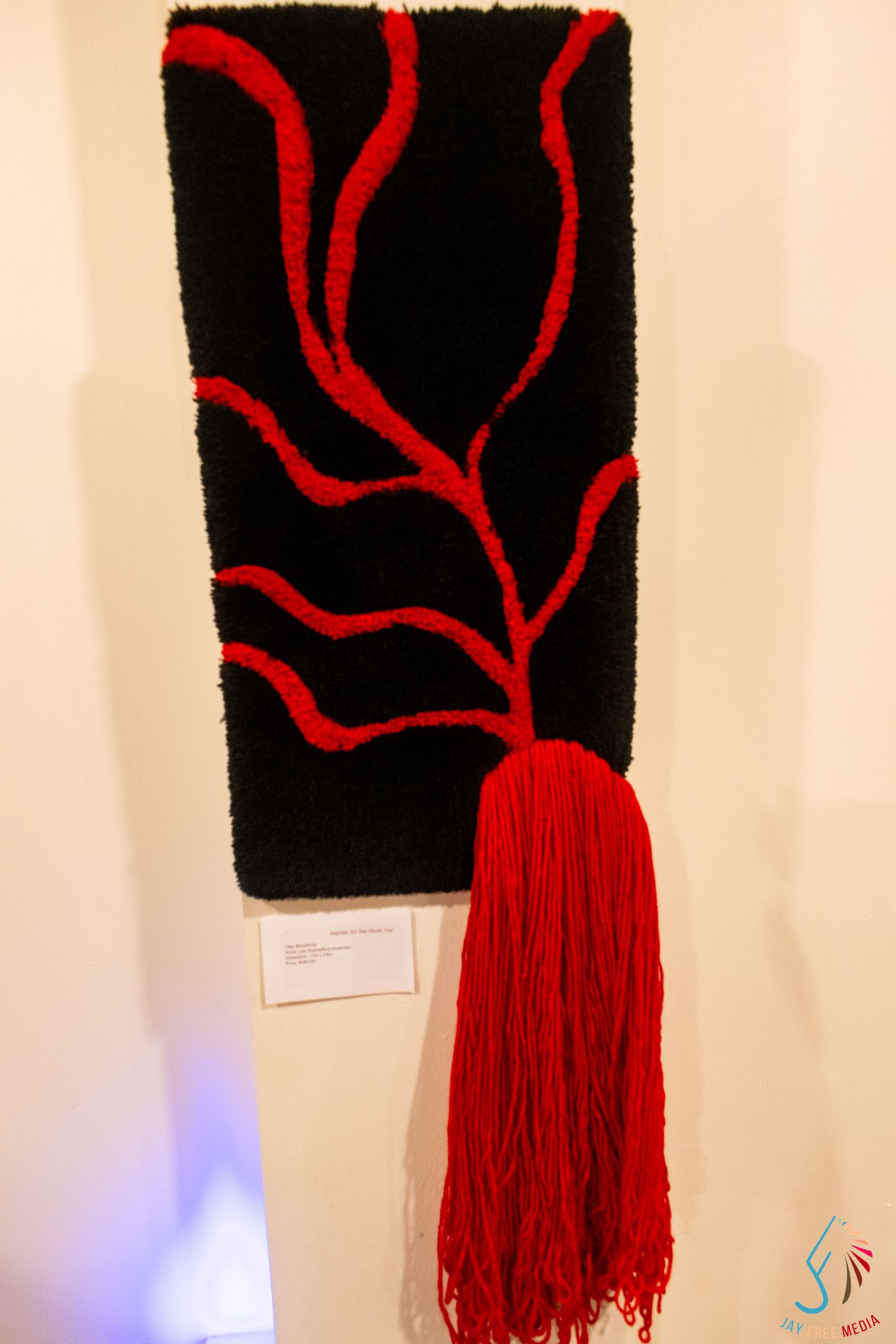Black textile wall art with red branching tree design and flowing red fringe tassel displayed on a white gallery wall.