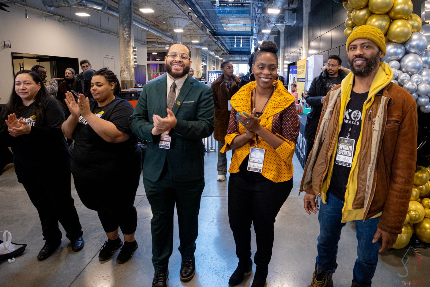 Diverse group of smiling people clapping and celebrating at an indoor event, dressed in colorful attire including yellow and green.