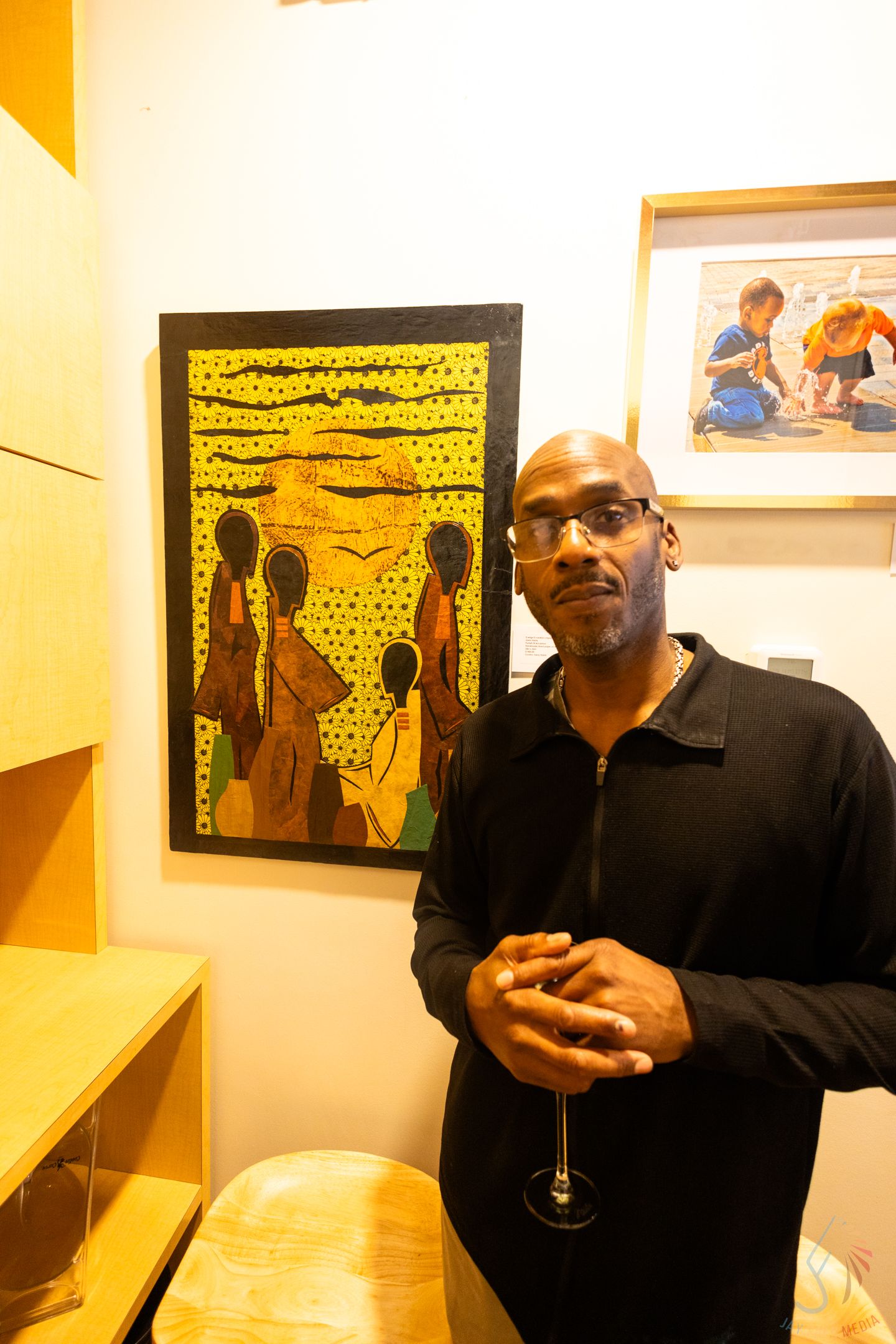 Man in black sweater holding wine glass poses beside colorful African art painting at gallery event.