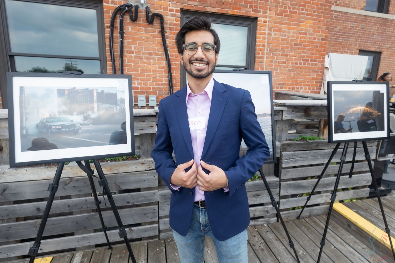 Man in blue blazer smiling outdoors at an art exhibition with framed photos on easels.