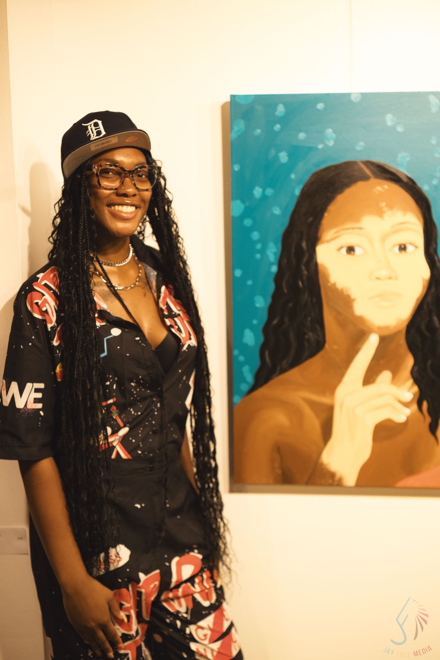 Woman with long braids in printed outfit stands next to portrait painting at gallery.
