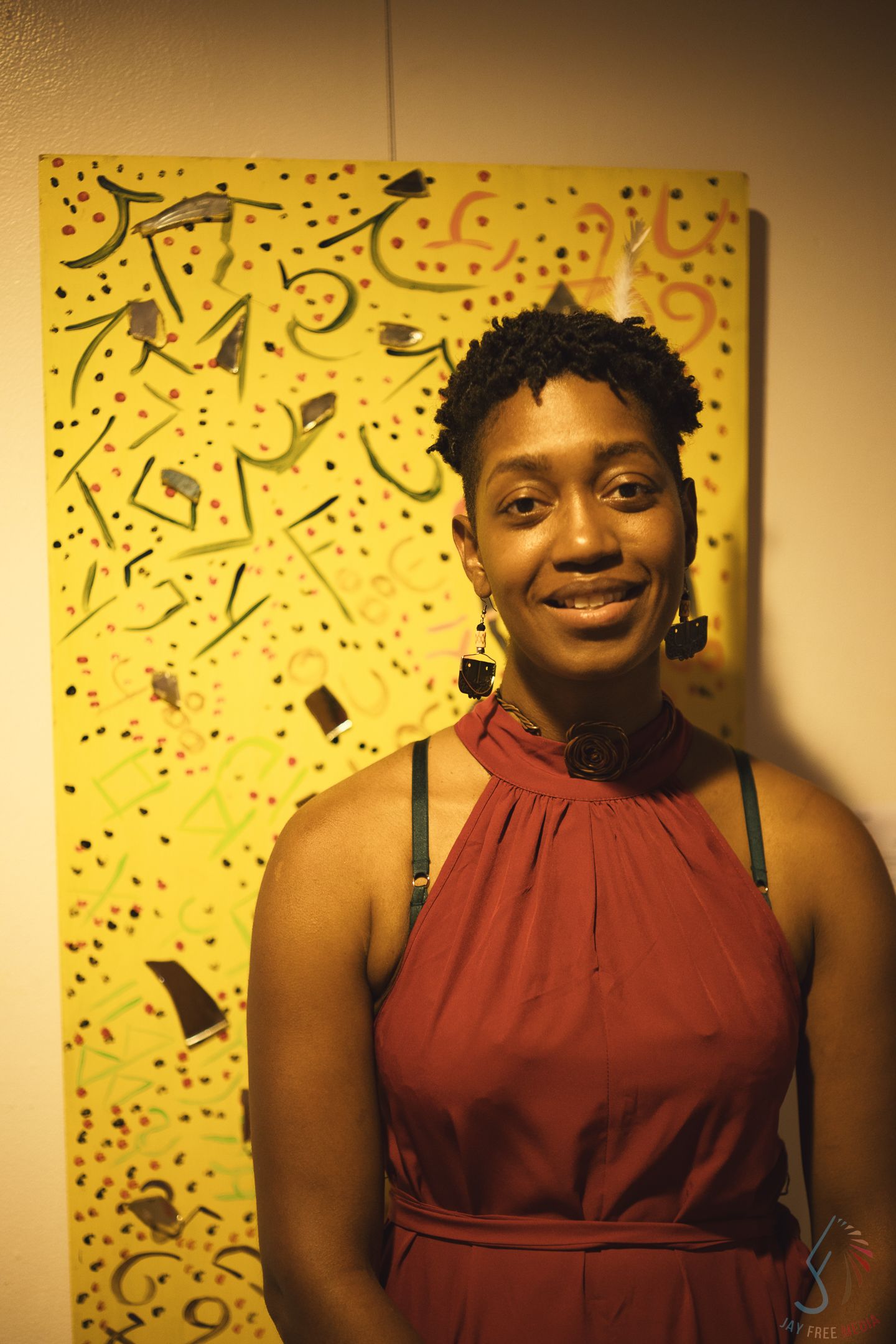 A smiling woman in a red halter dress poses in front of a vibrant yellow abstract painting at an art gallery.