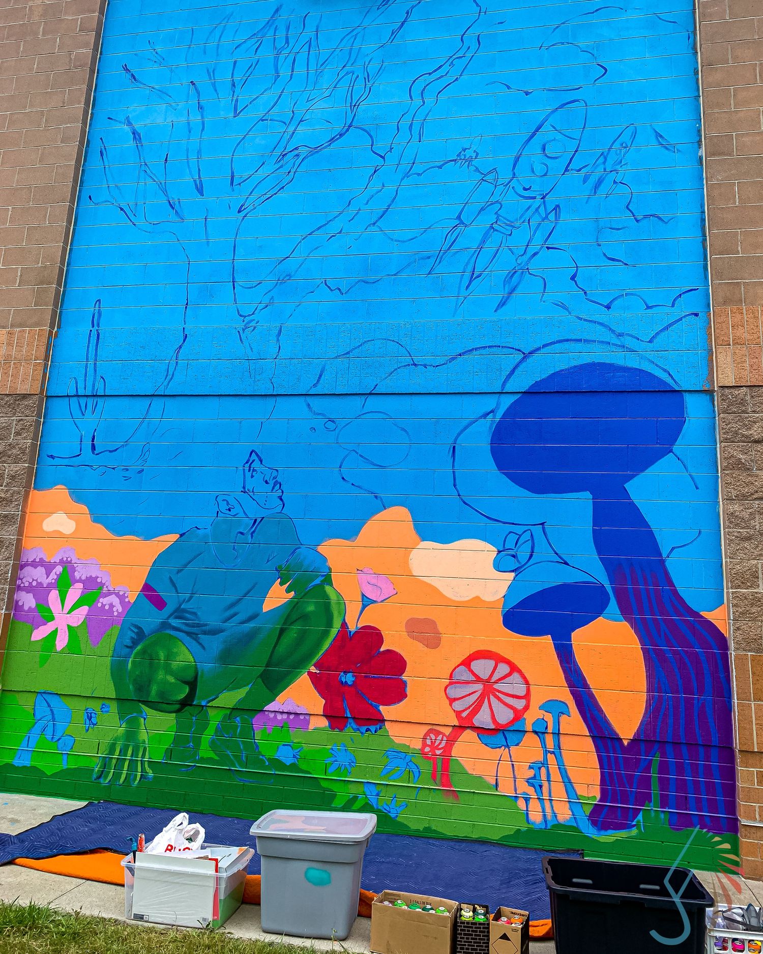 Colorful mural in progress on brick wall featuring flowers, mushrooms, and figures in vibrant blues, greens, and oranges.