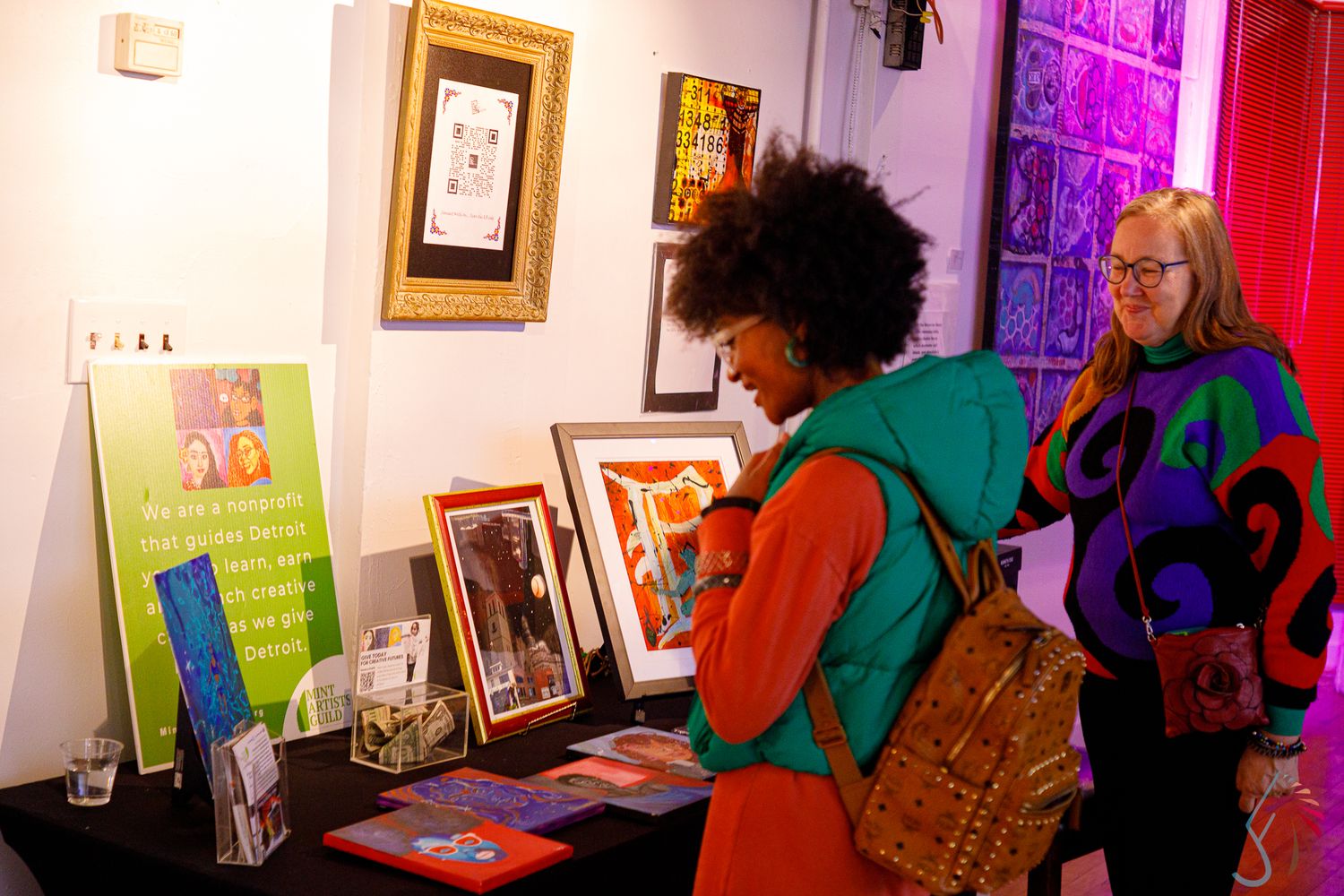 Two women browse colorful artwork and framed prints displayed on a table at an indoor art gallery exhibition.