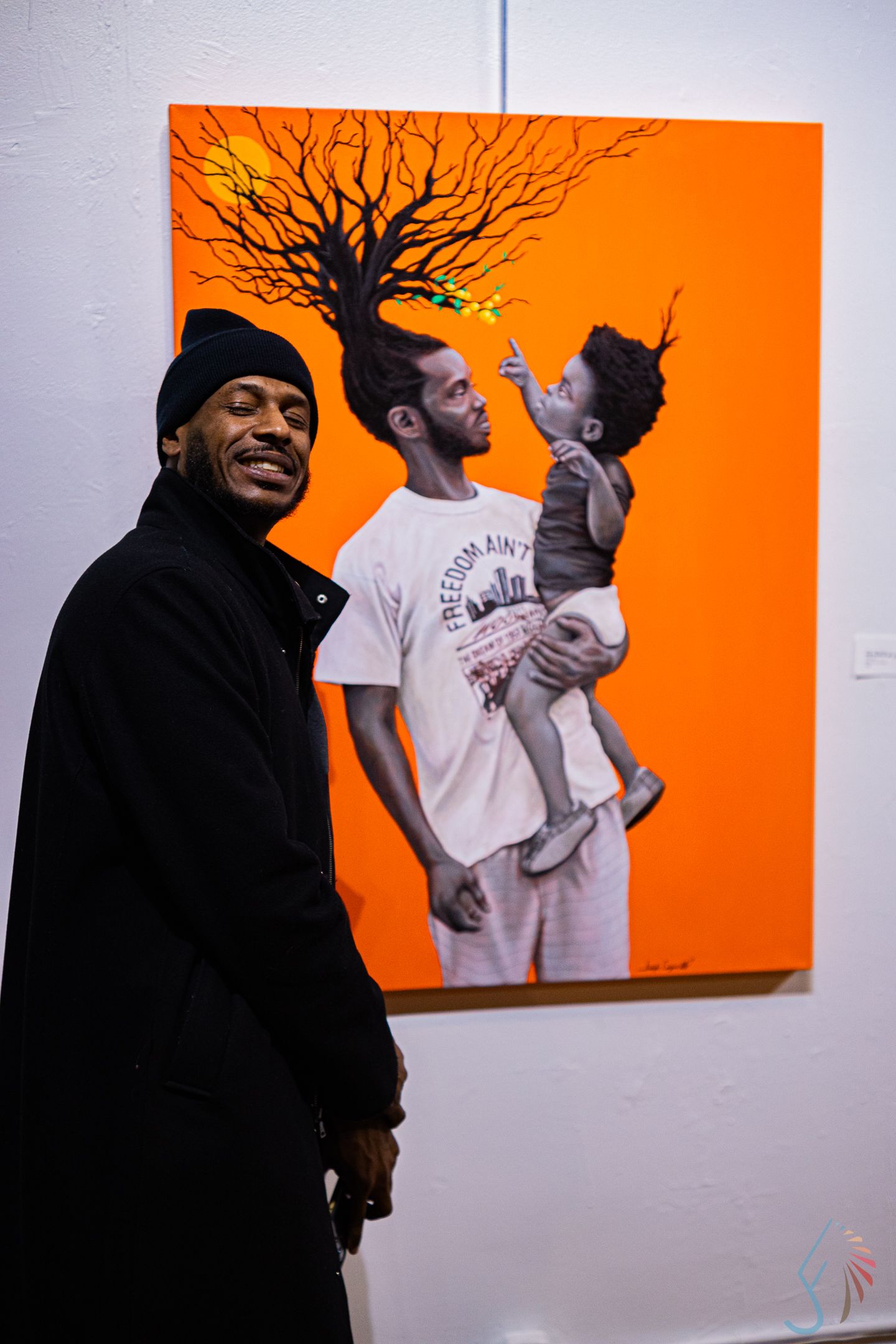 Man in black outfit smiles beside orange painting of father and child at gallery.