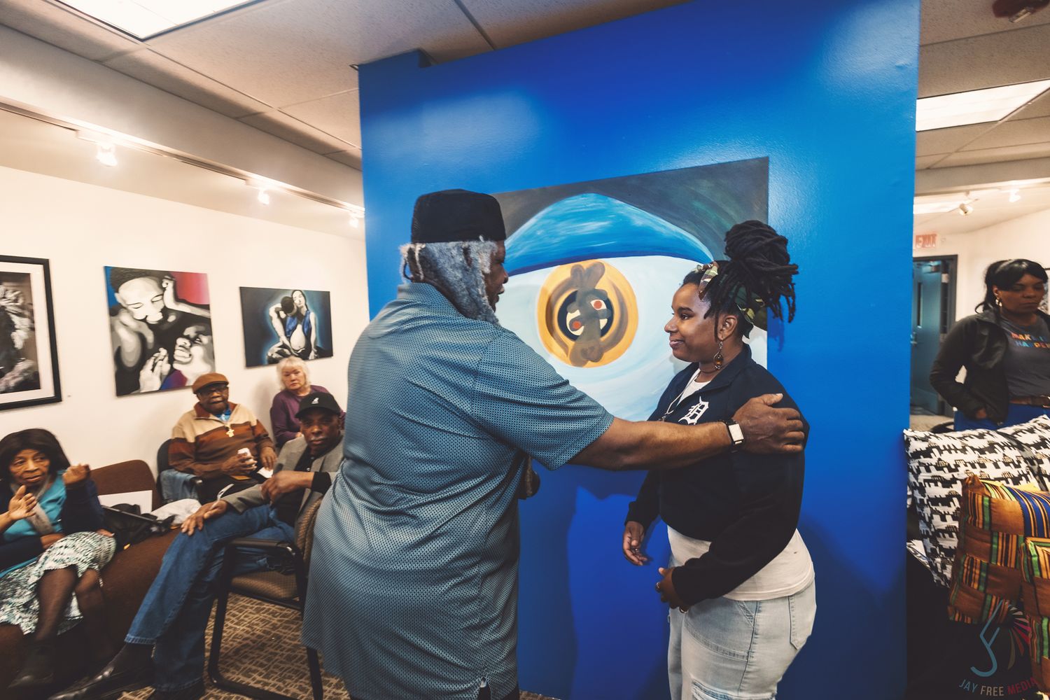 Two people embracing in a vibrant blue art gallery with diverse artwork on the walls and seated guests nearby.