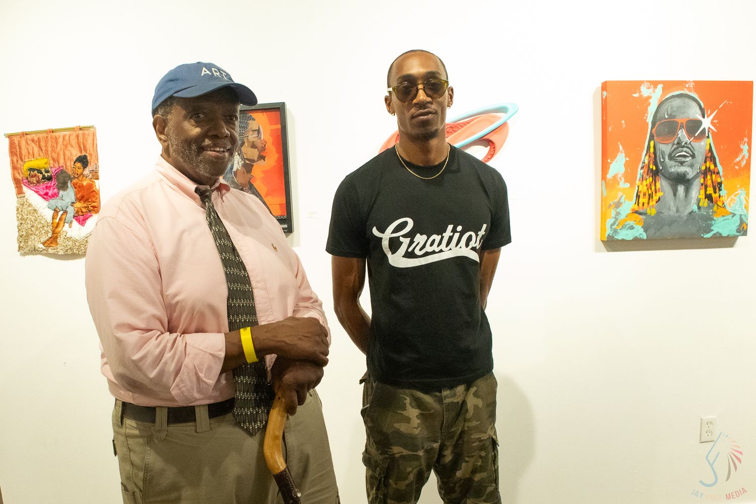 Two men posing at an art gallery with colorful paintings displayed on the white walls behind them.