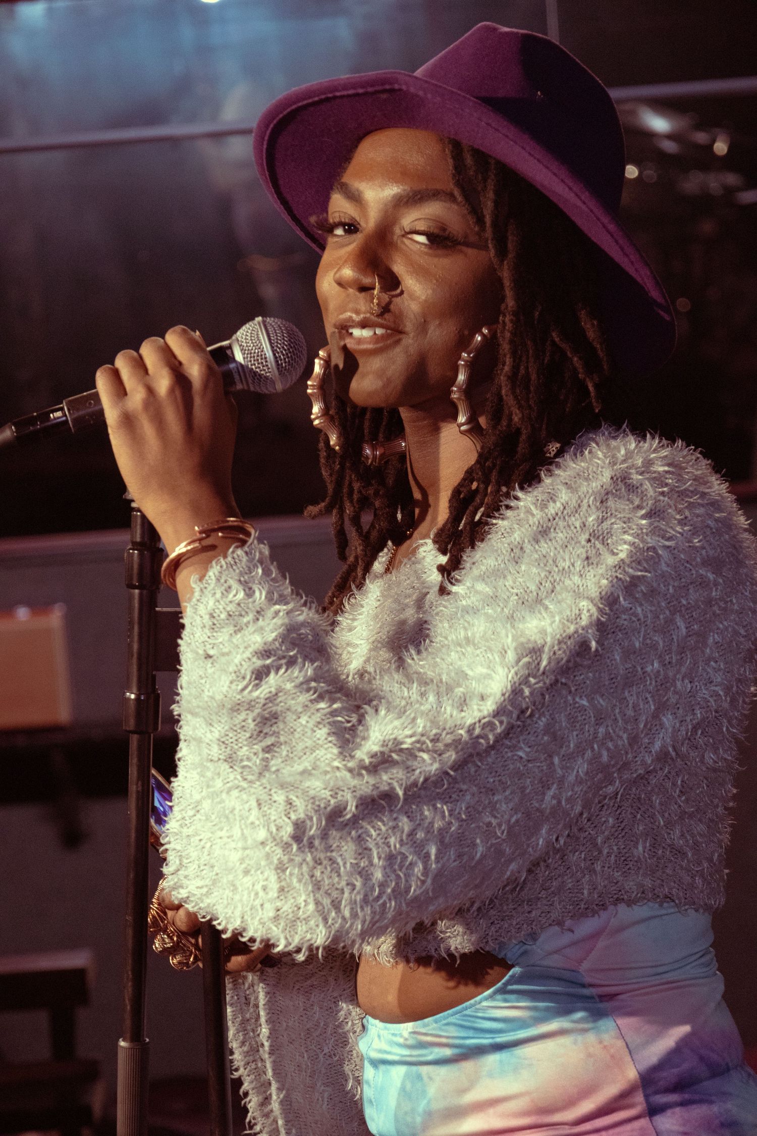 A young Black woman singer performs on stage wearing a purple hat and fluffy gray jacket while holding a microphone.