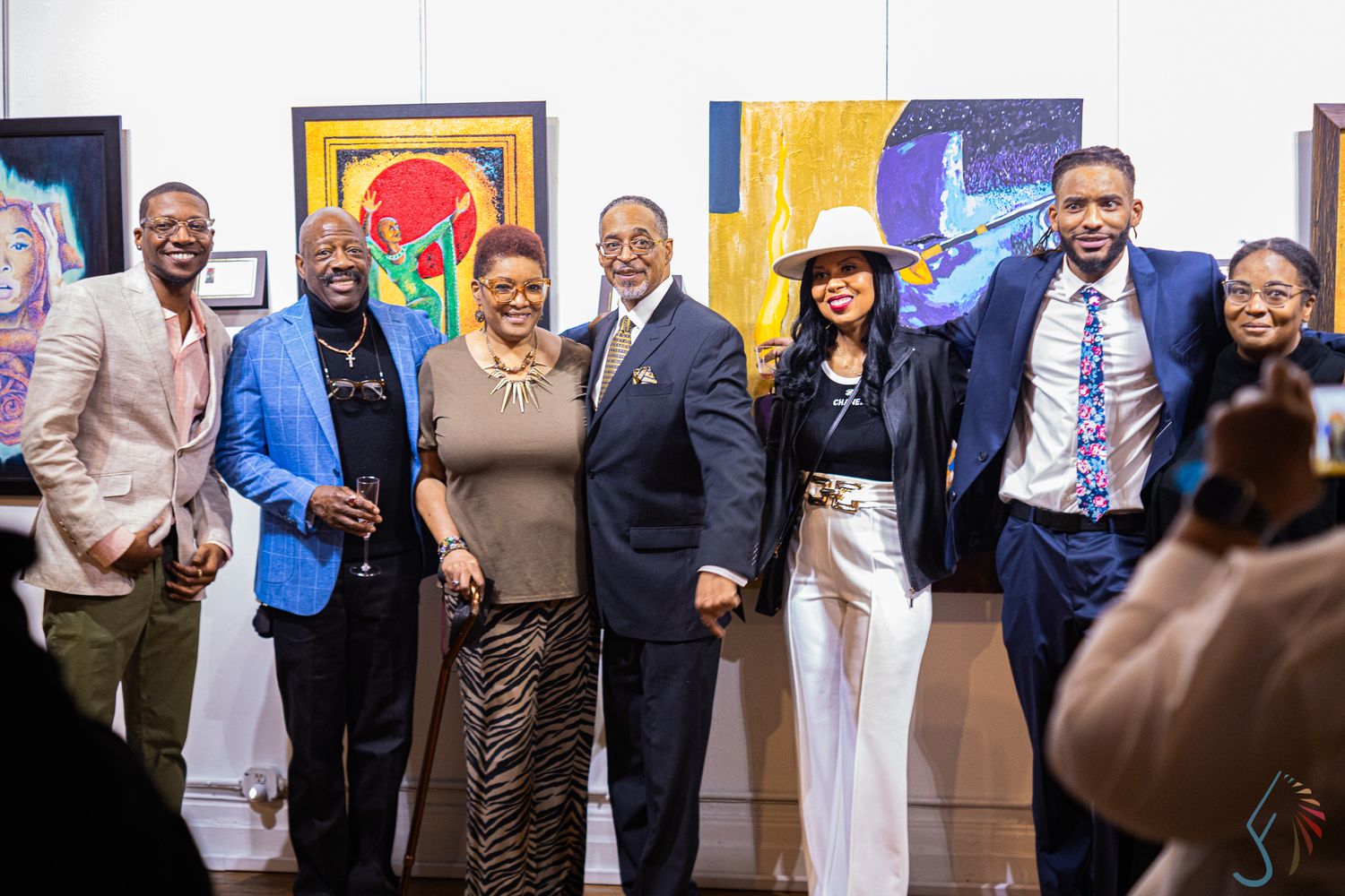 Group of seven people posing together at an art gallery event with colorful paintings displayed on the wall behind them.