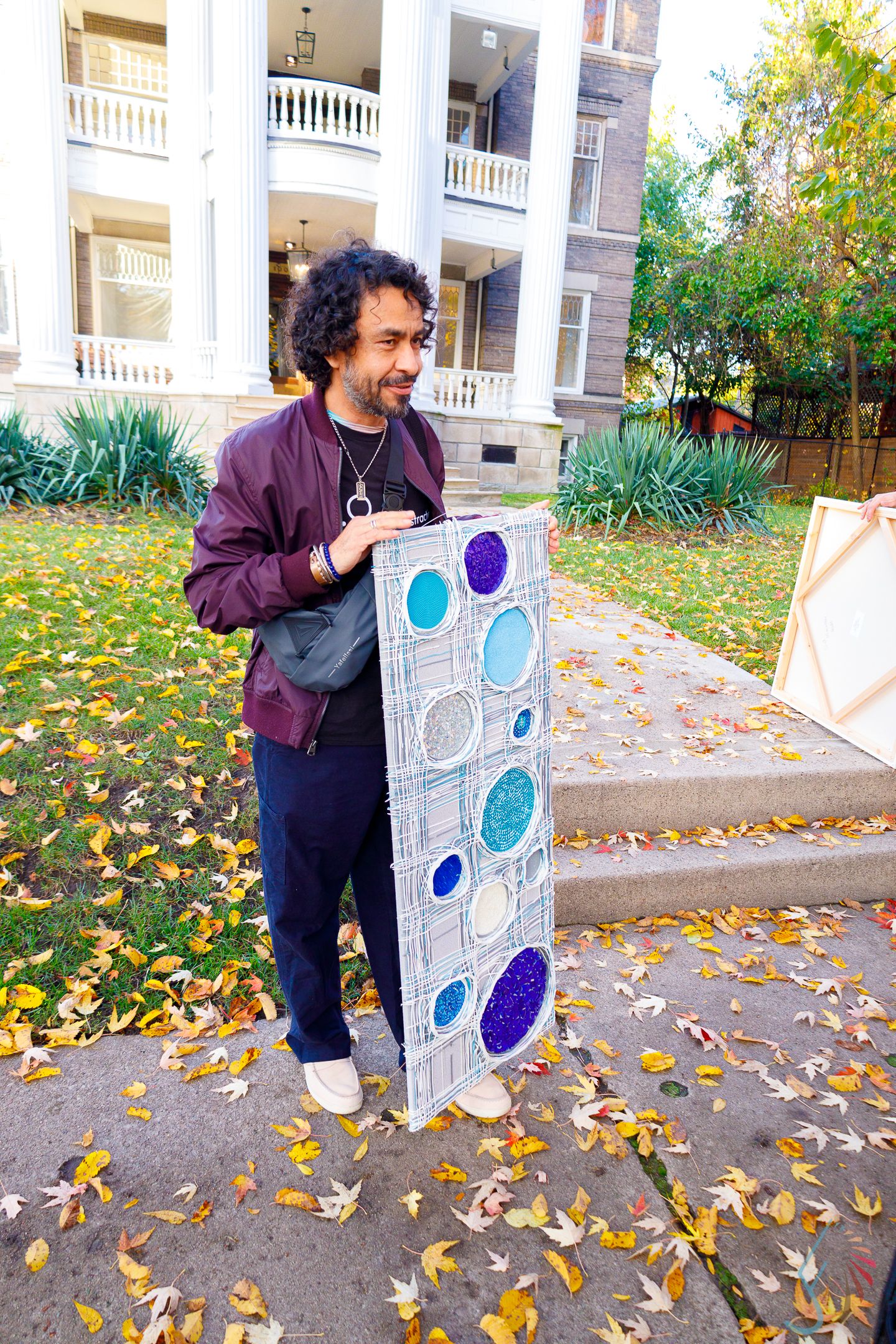 Man in purple jacket holds colorful circular patterned artwork outdoors among autumn leaves near historic white building.