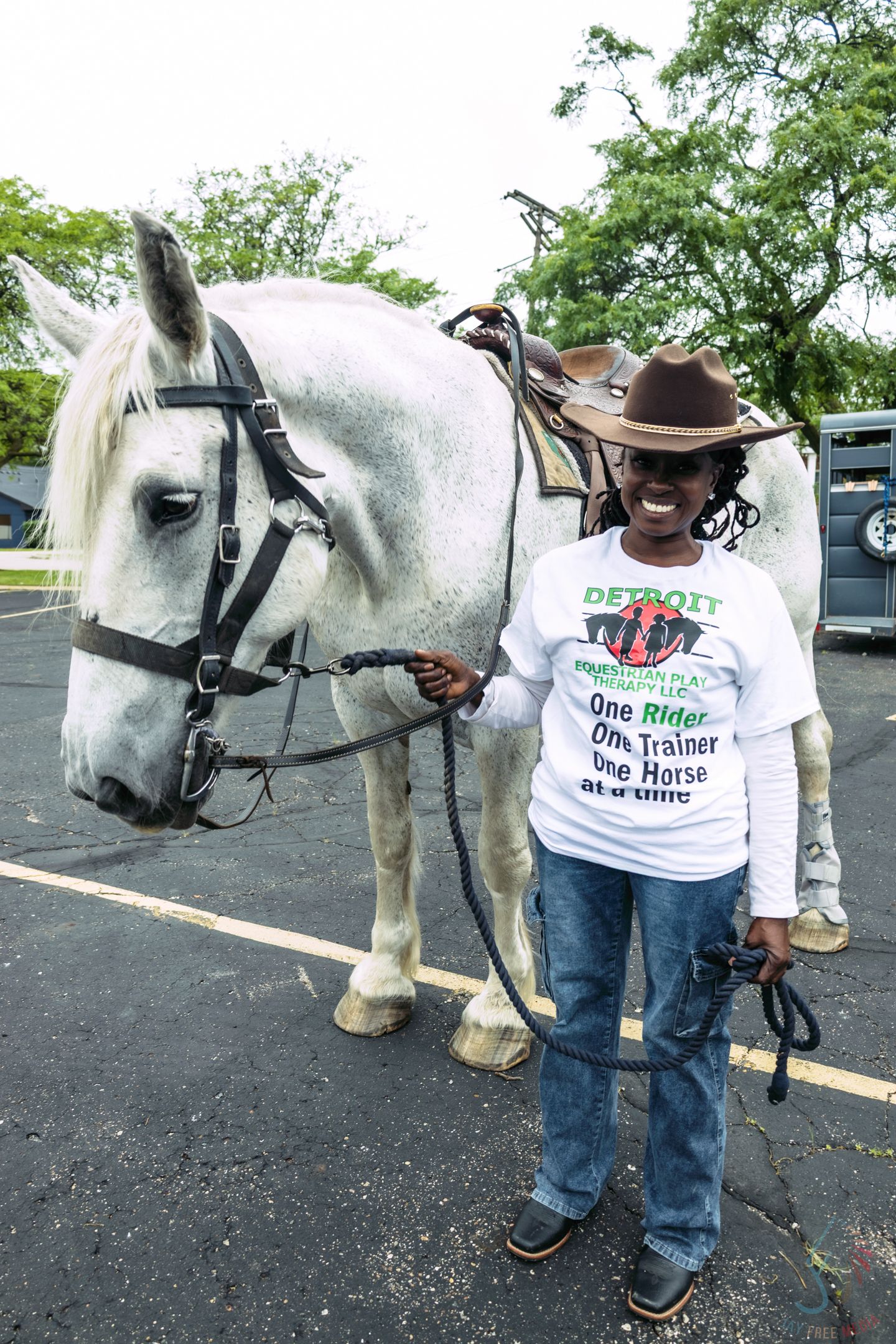 Smiling person in Detroit shirt and cowboy hat stands beside a white horse in a parking lot.