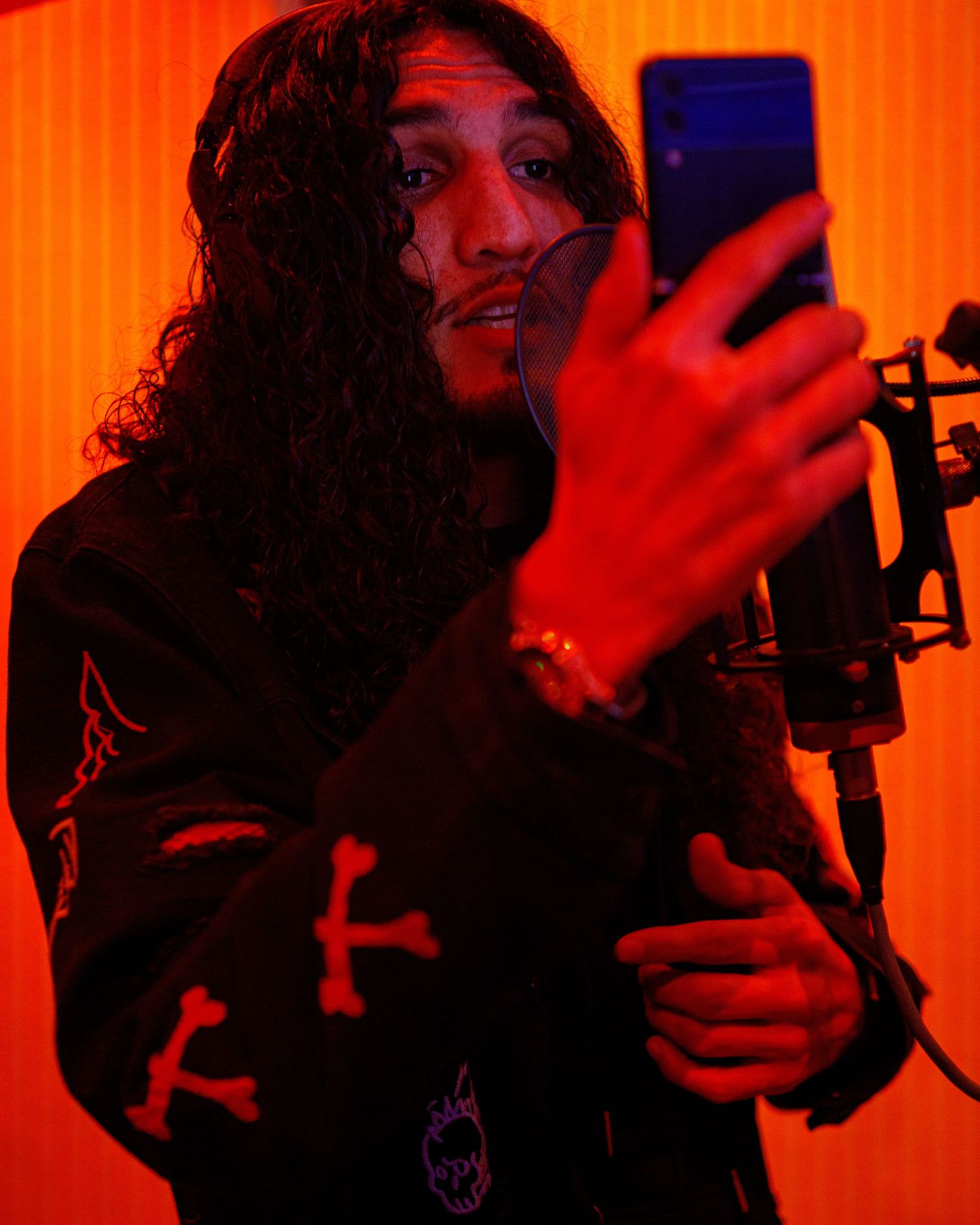 Musician with long curly hair recording in studio with microphone and phone.