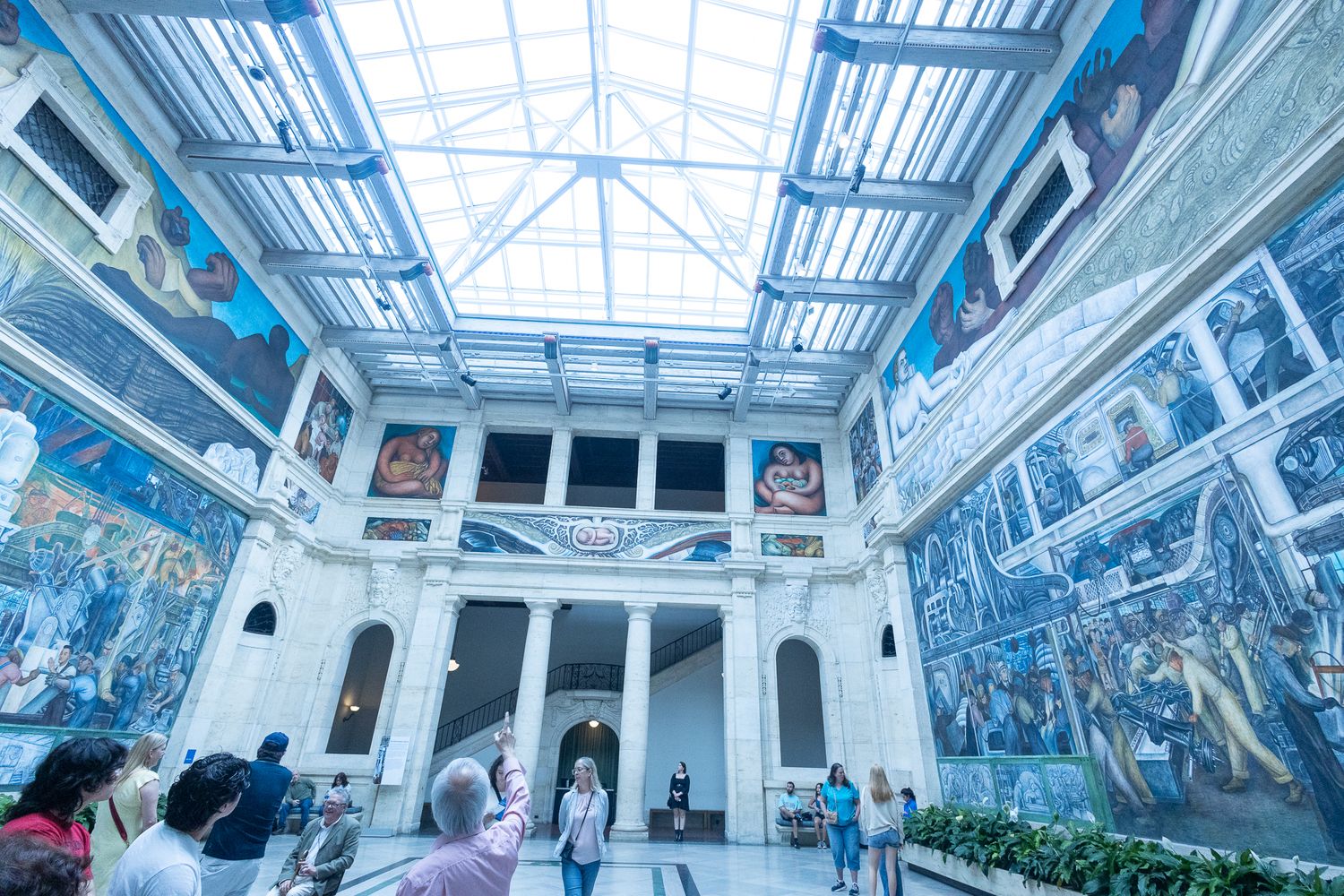 Grand museum hall with large-scale murals covering walls under a glass skylight ceiling with visitors.
