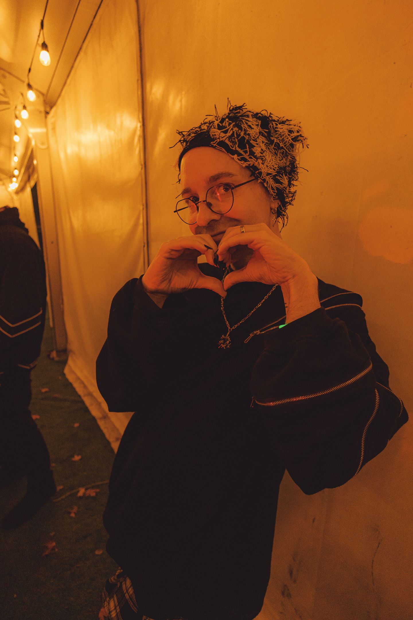 Person in black jacket and patterned headscarf making a heart shape with hands under warm string lights.