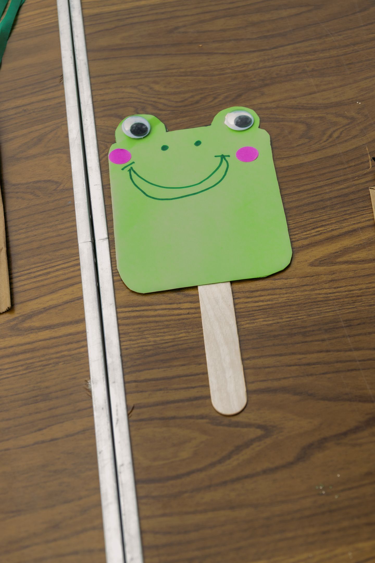 Handmade green frog puppet craft on a popsicle stick with googly eyes on a wooden table.