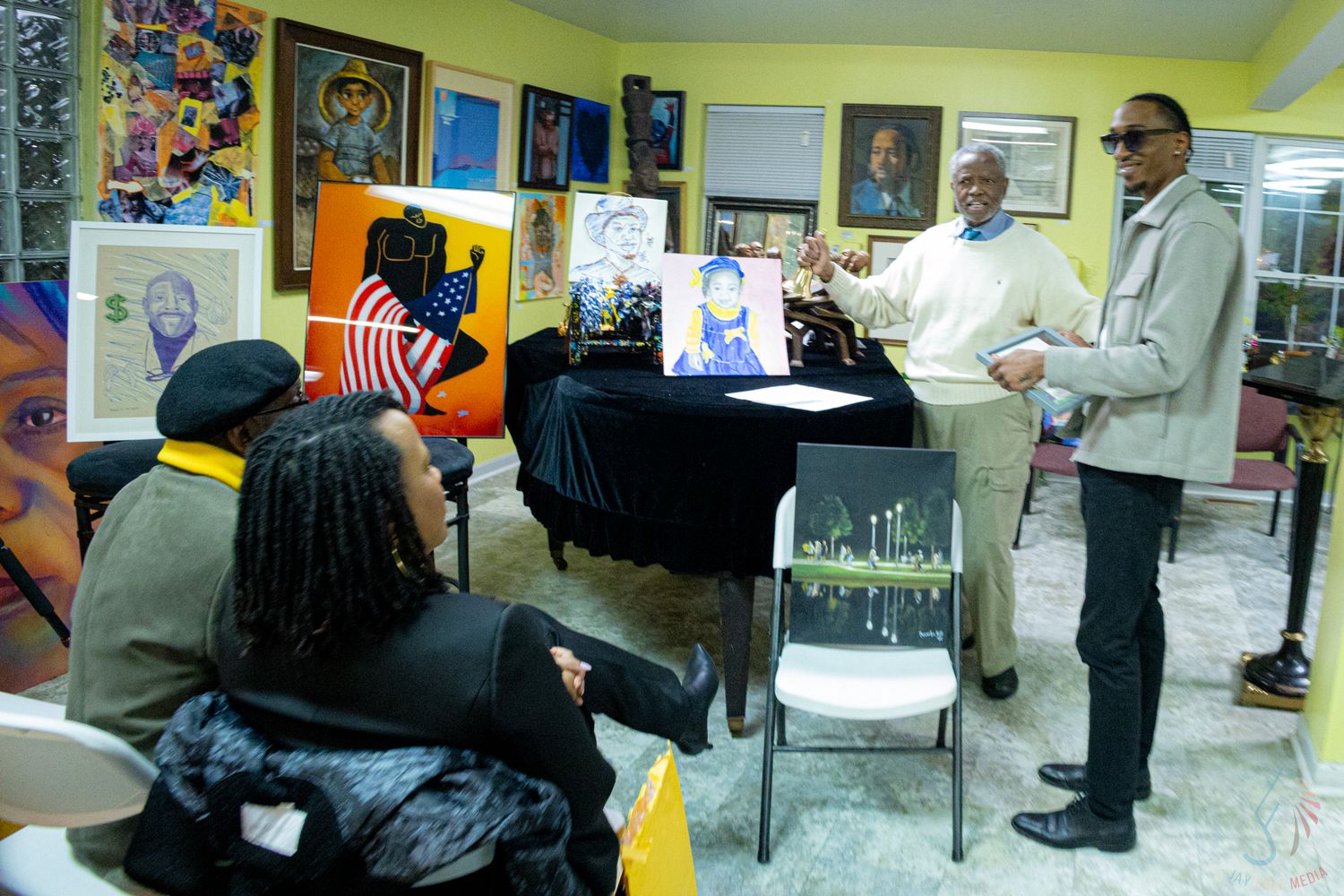 People gathered in an art gallery for a community meeting, with colorful paintings displayed on yellow walls.
