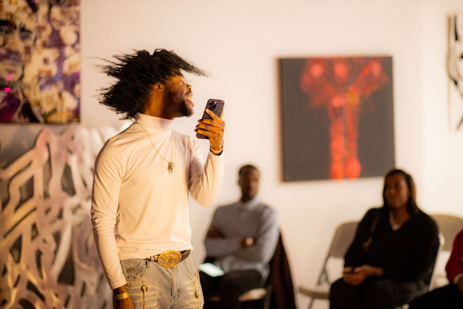 A man with voluminous natural hair reads from his phone while performing spoken word at an art gallery event.