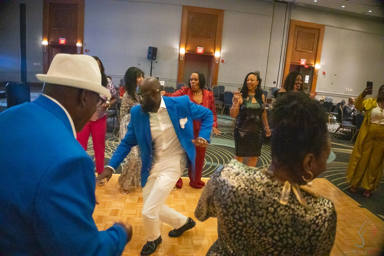 Man in blue blazer and white hat energetically dancing at a formal ballroom event.