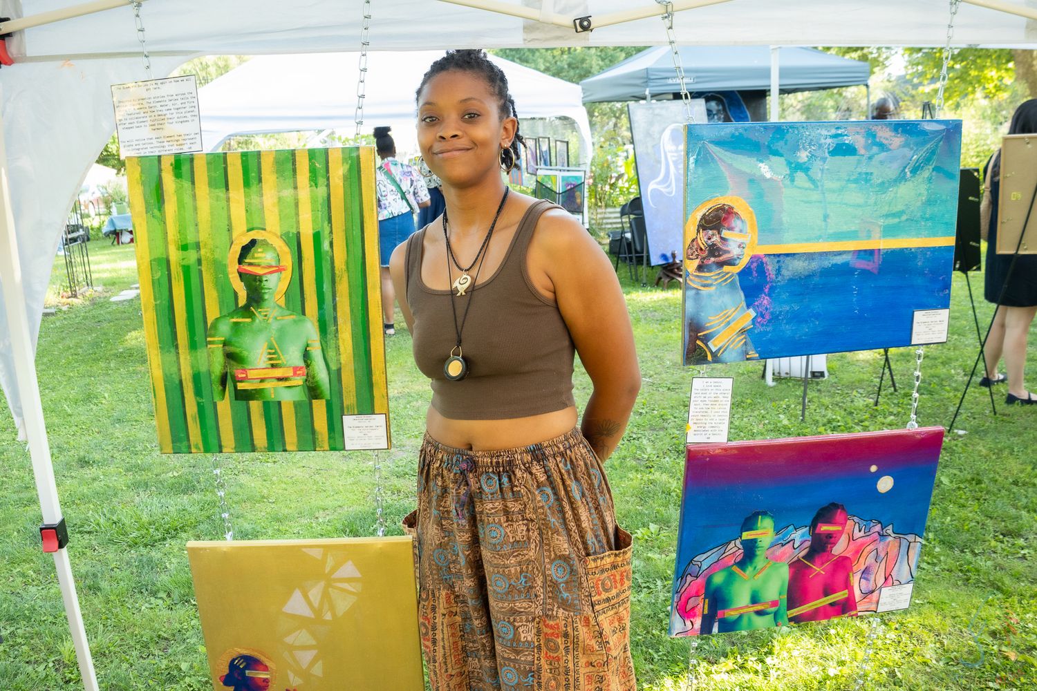 Young woman poses proudly at her outdoor art booth displaying colorful paintings under a white tent.