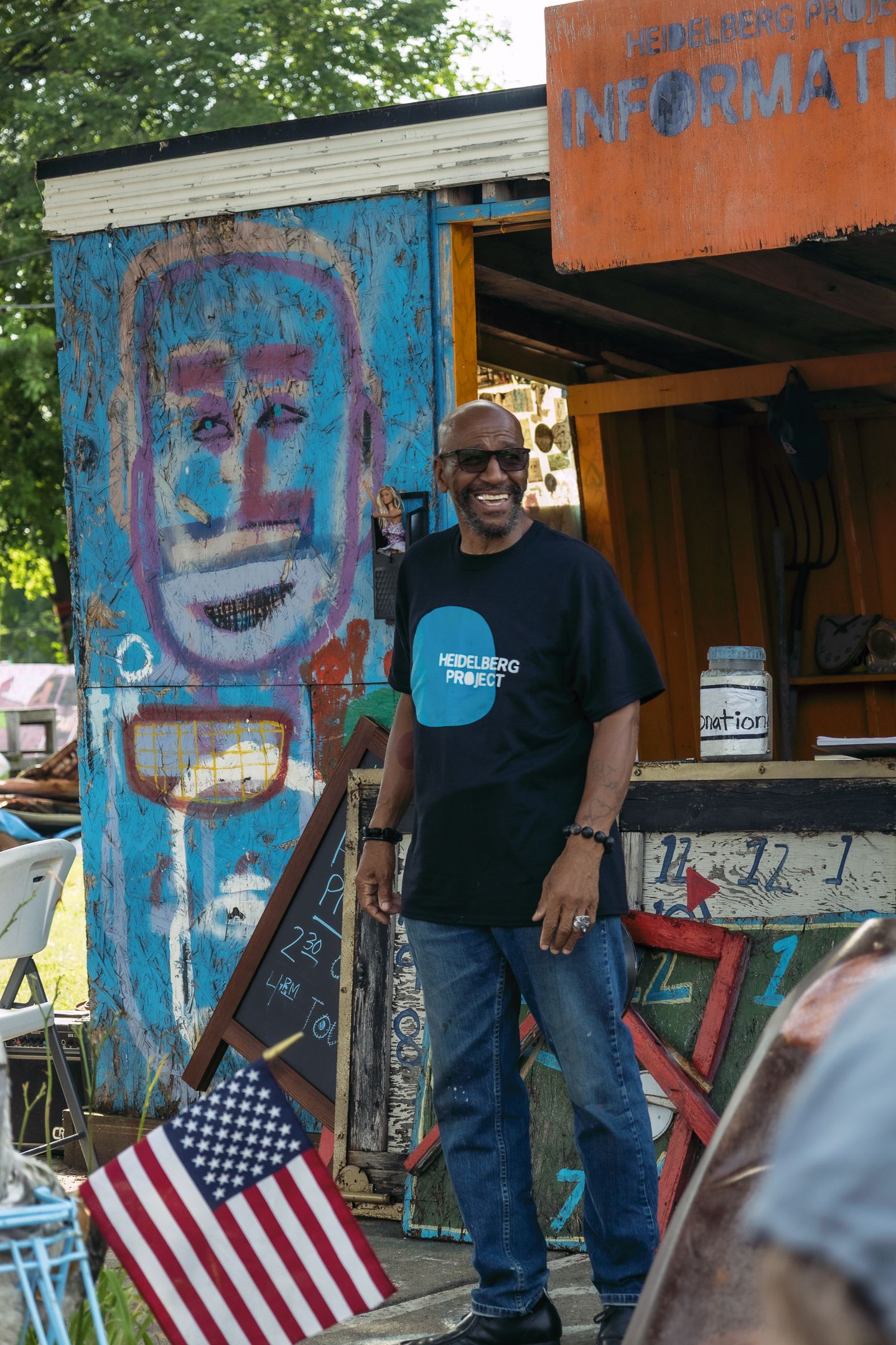 Man in Heidelberg Project t-shirt smiling outdoors near colorful mural art and American flag in Detroit.
