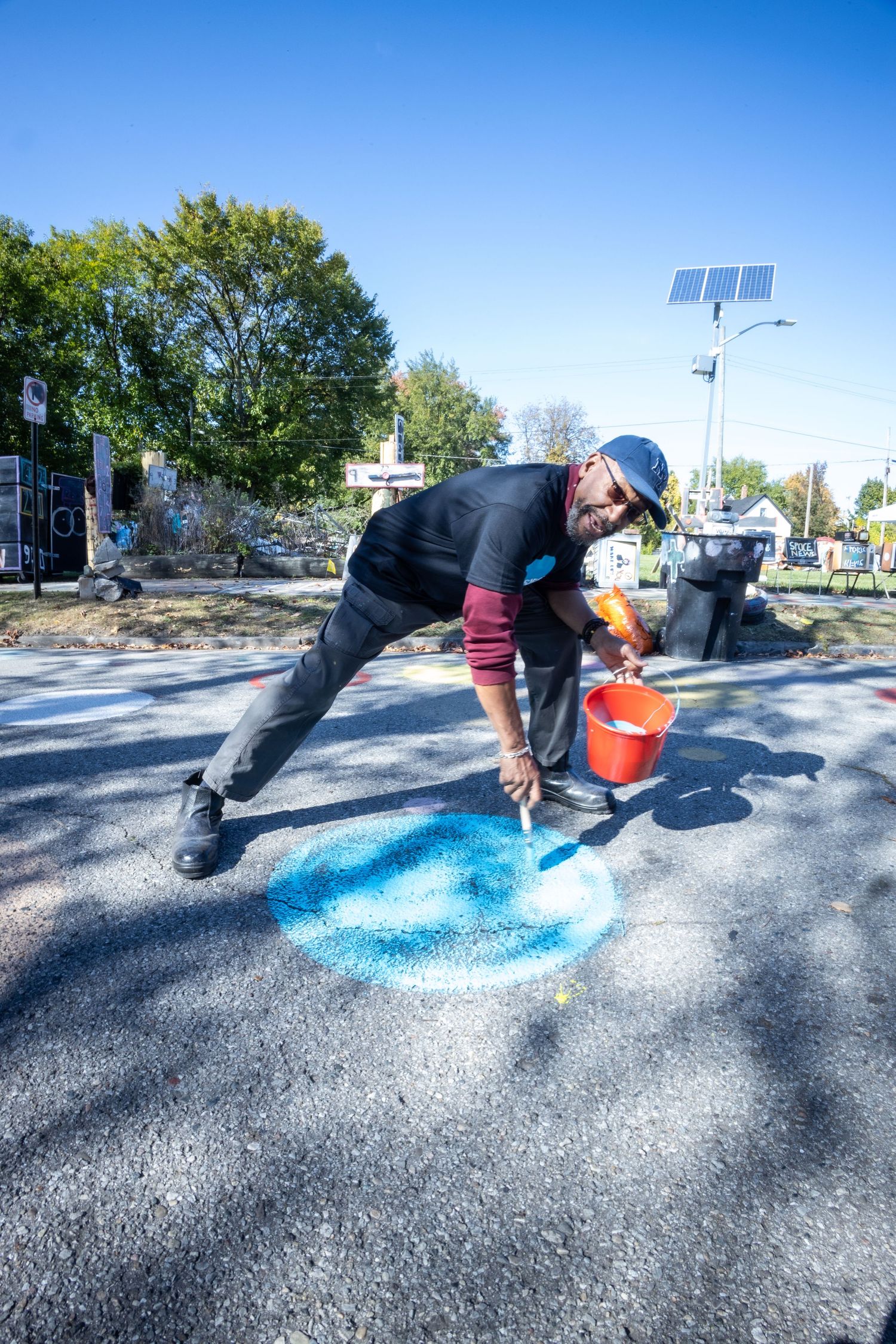 Artist leaning over to paint a blue circle on asphalt pavement with a red bucket during a street art project.