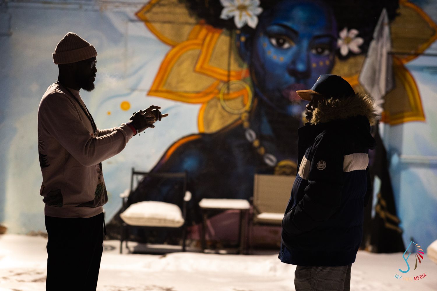 Two people conversing in front of a vibrant blue goddess mural painted on a wall in an urban setting.