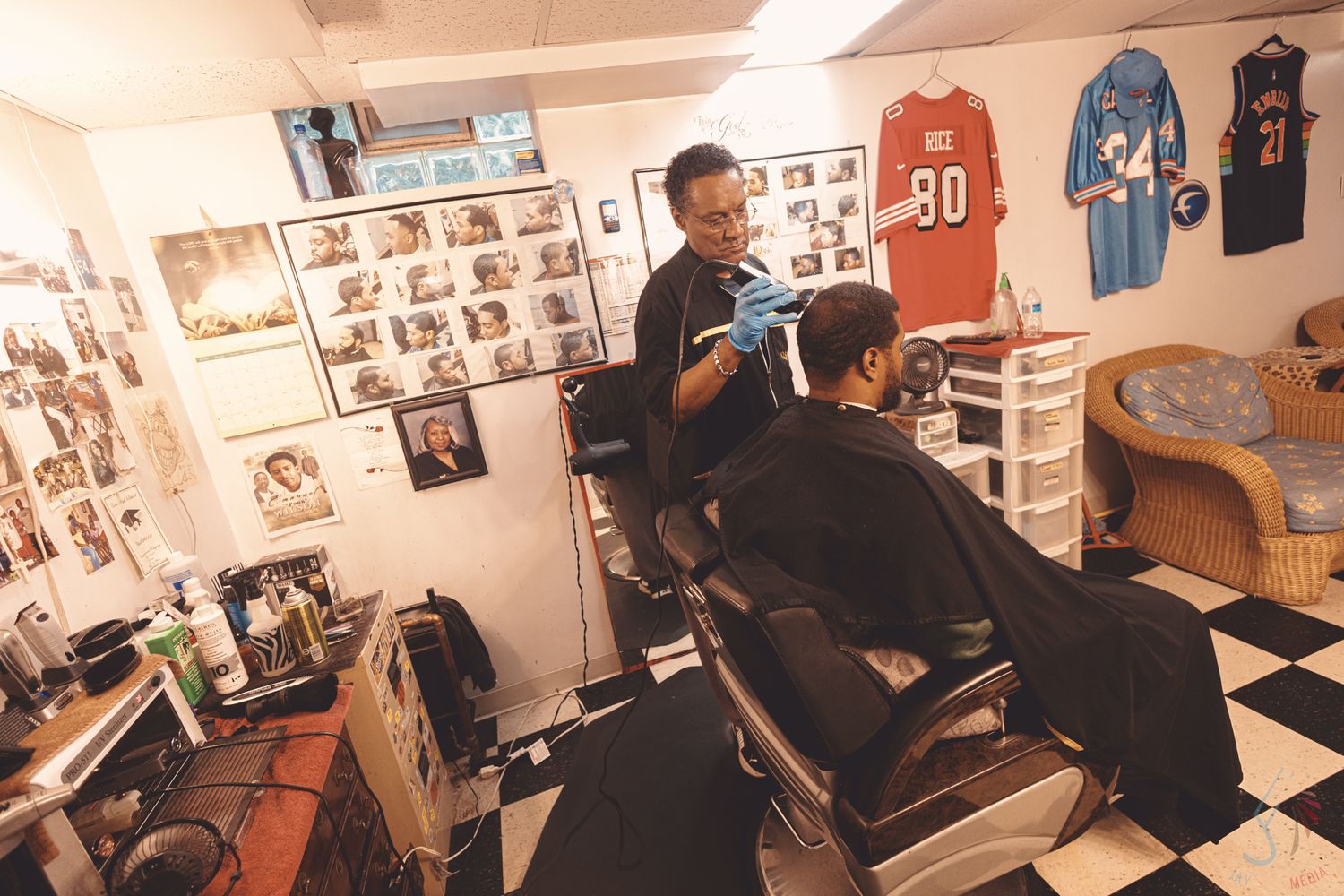 Barber cutting a client's hair in a decorated shop with sports jerseys and black-and-white photos on the walls.