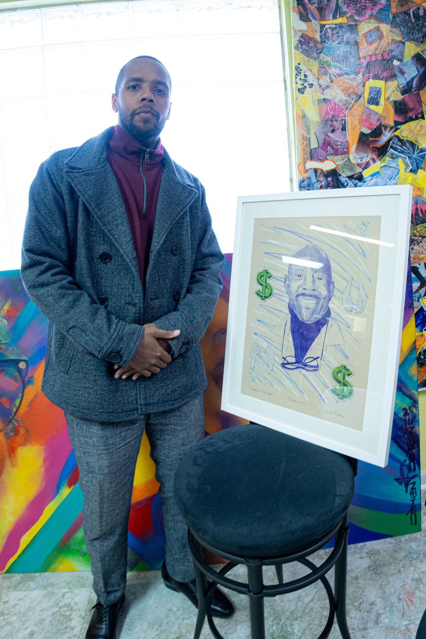 Man in gray coat stands beside framed portrait artwork with dollar signs, surrounded by colorful paintings in gallery.