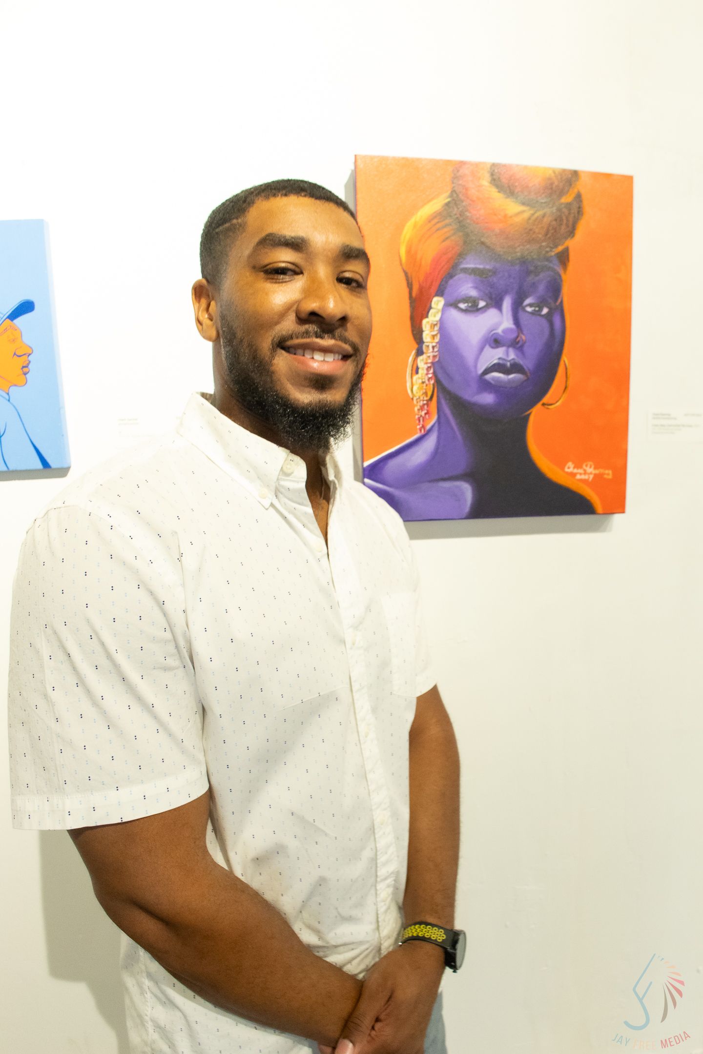 Man in white shirt smiling at art gallery with colorful portrait paintings on wall behind him.